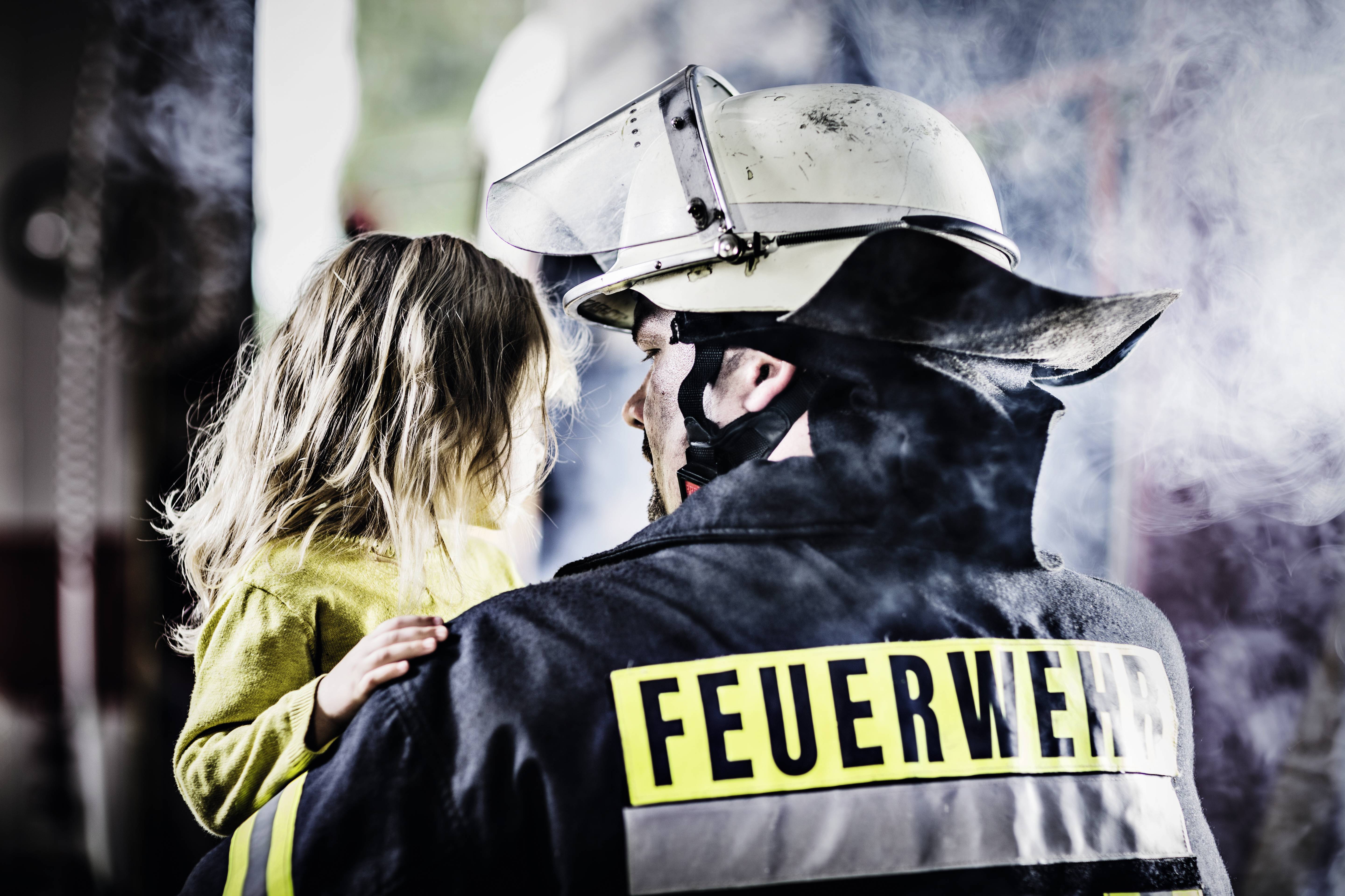 Brandweerman in uniform draagt een klein meisje door een rookruimte. Ze kijken elkaar aan, waarbij het meisje zich stevig vasthoudt.