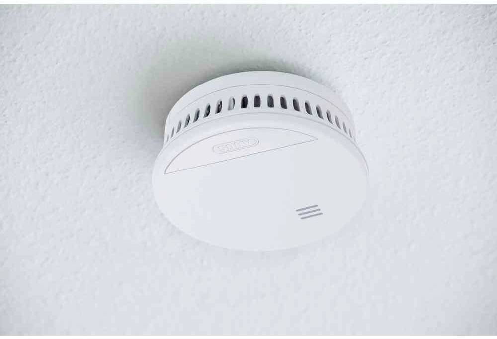 Een witte rookmelder is aan het plafond bevestigd. Hij heeft verluchtingssleuven aan de zijkant en een eenvoudig, rond ontwerp.