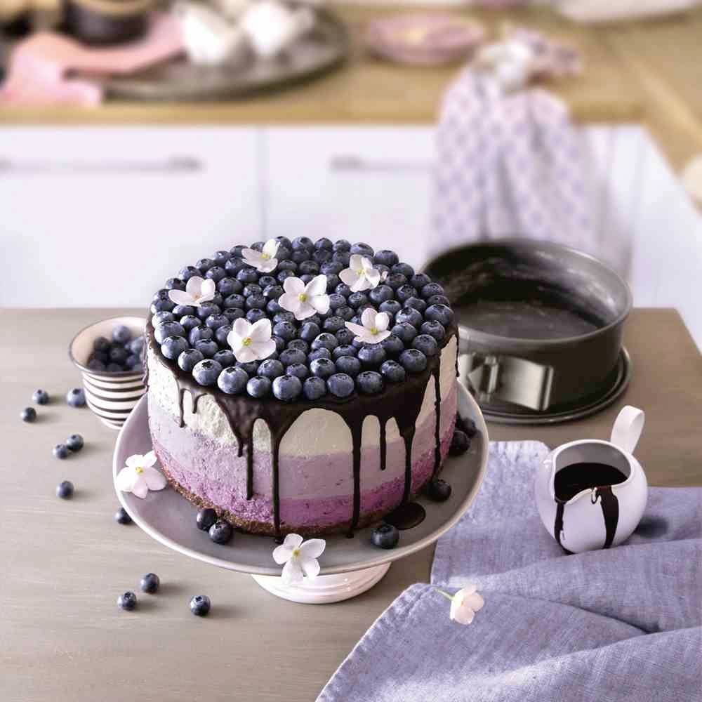 Een drielaagde taart met chocoladeglazuur, versierd met bosbessen en witte bloemen op een tafel in een keuken.