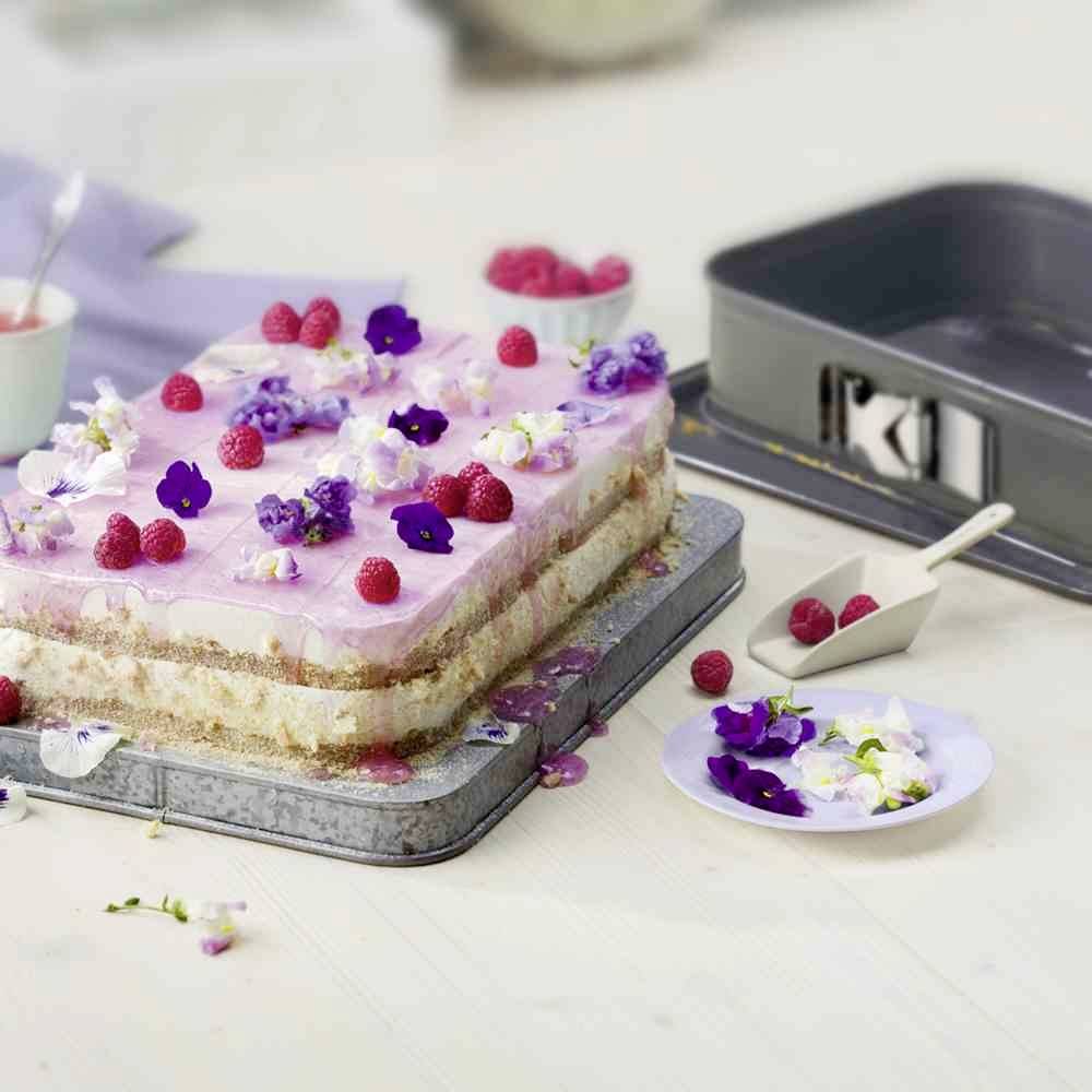 Een tweeLaagse taart met roze glazuur, gegarneerd met frambozen en eetbare paarse bloemen op een metalen bakplaat.