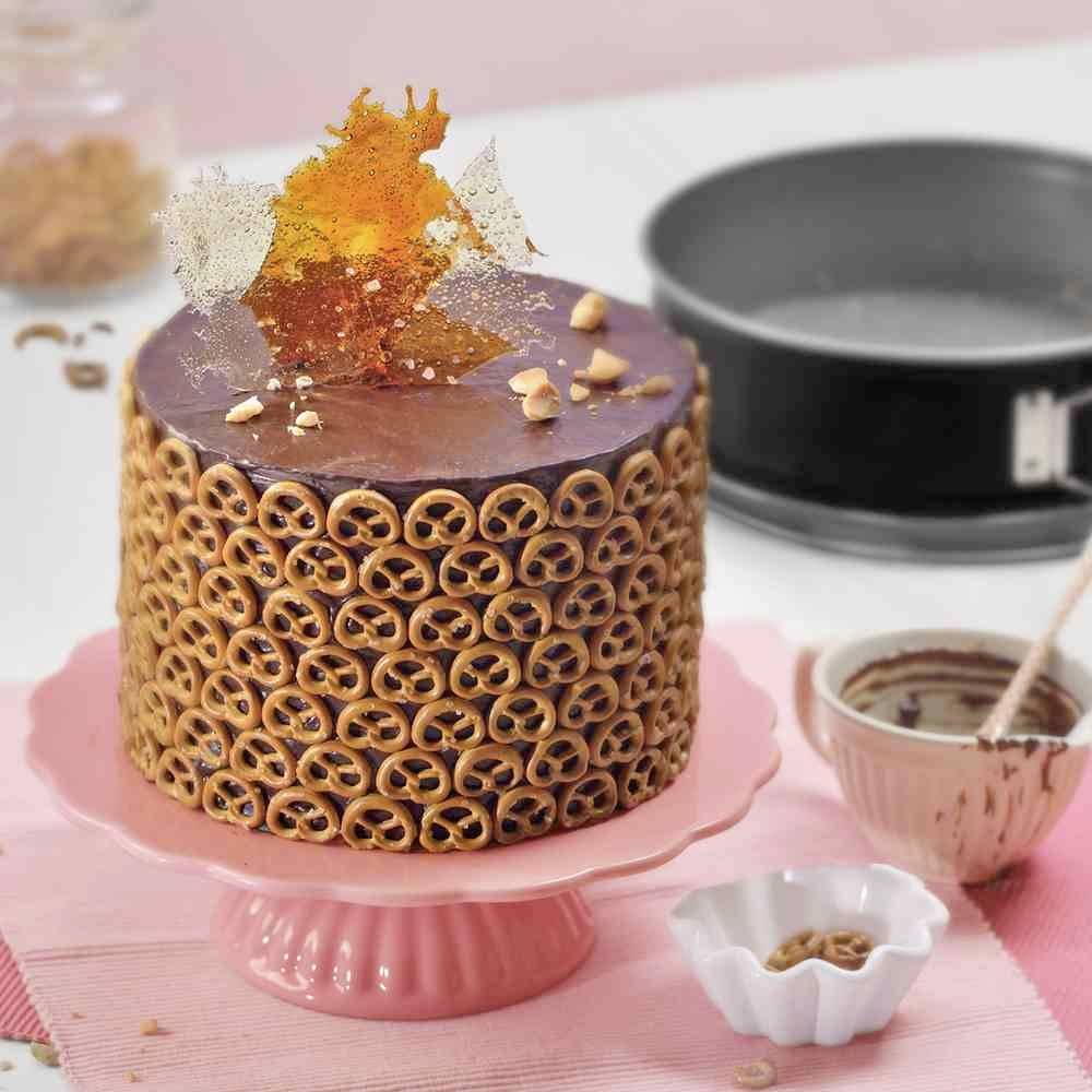 Een ronde taart bedekt met zoute pretzels, versierd met karamelfragmenten, staat op een roze taartschaal. Op de achtergrond liggen bakbenodigdheden.