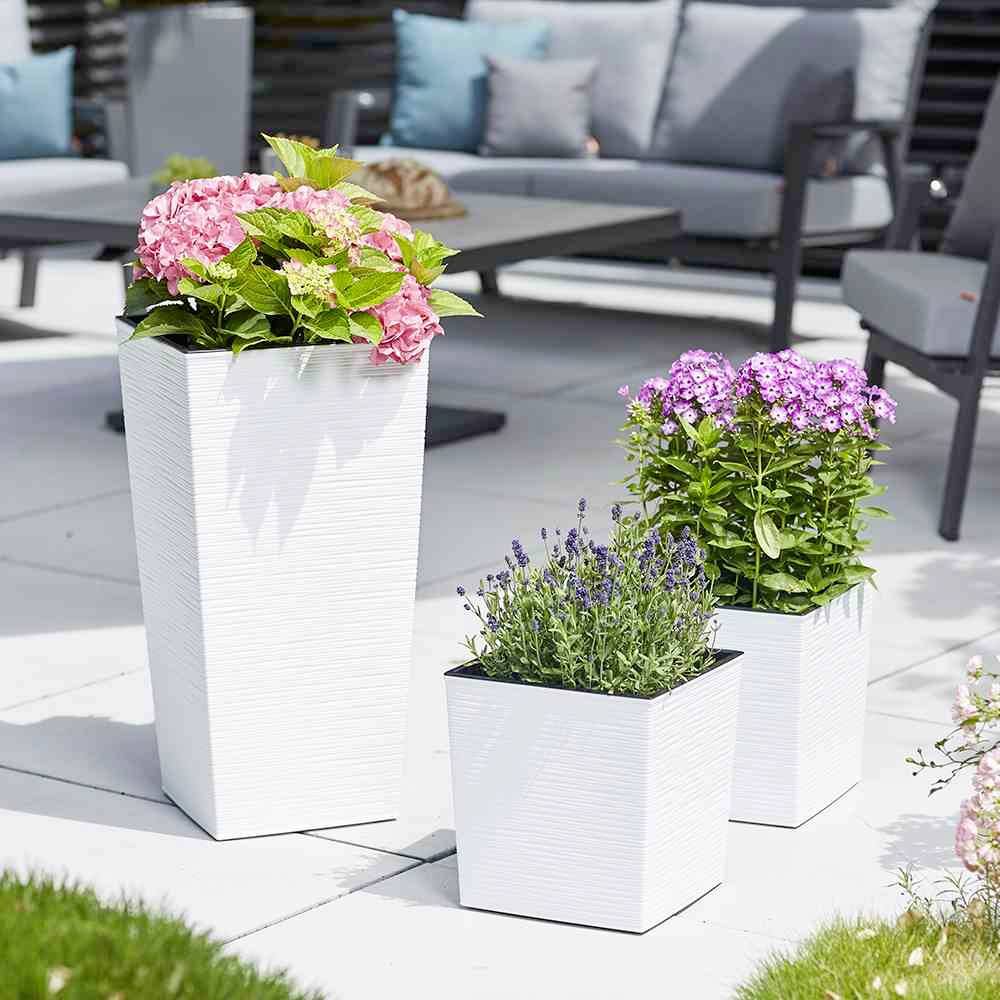 Drie witte bloempotten met roze hortensia's, lavendel en paarse bloemen op een terras. Op de achtergrond tuinmeubels.
