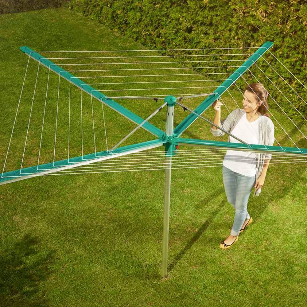 Een vrouw staat in de tuin en bedient een groene droogmolen met uitgespreide lijnen, gebruikt om was te drogen.