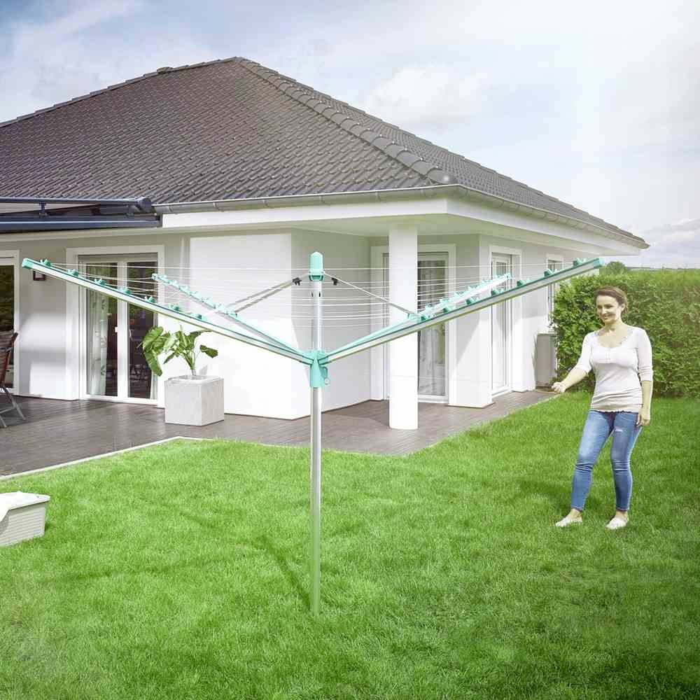 Een vrouw staat in de tuin naast een opengevouwen droogrek op groen gazon voor een wit huis met een terras.