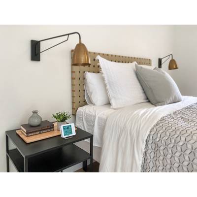 Een eenvoudige slaapkamer met een versierd bed, twee wandlampen en een nachtkastje waarop boeken, een vaas en een wekker staan.