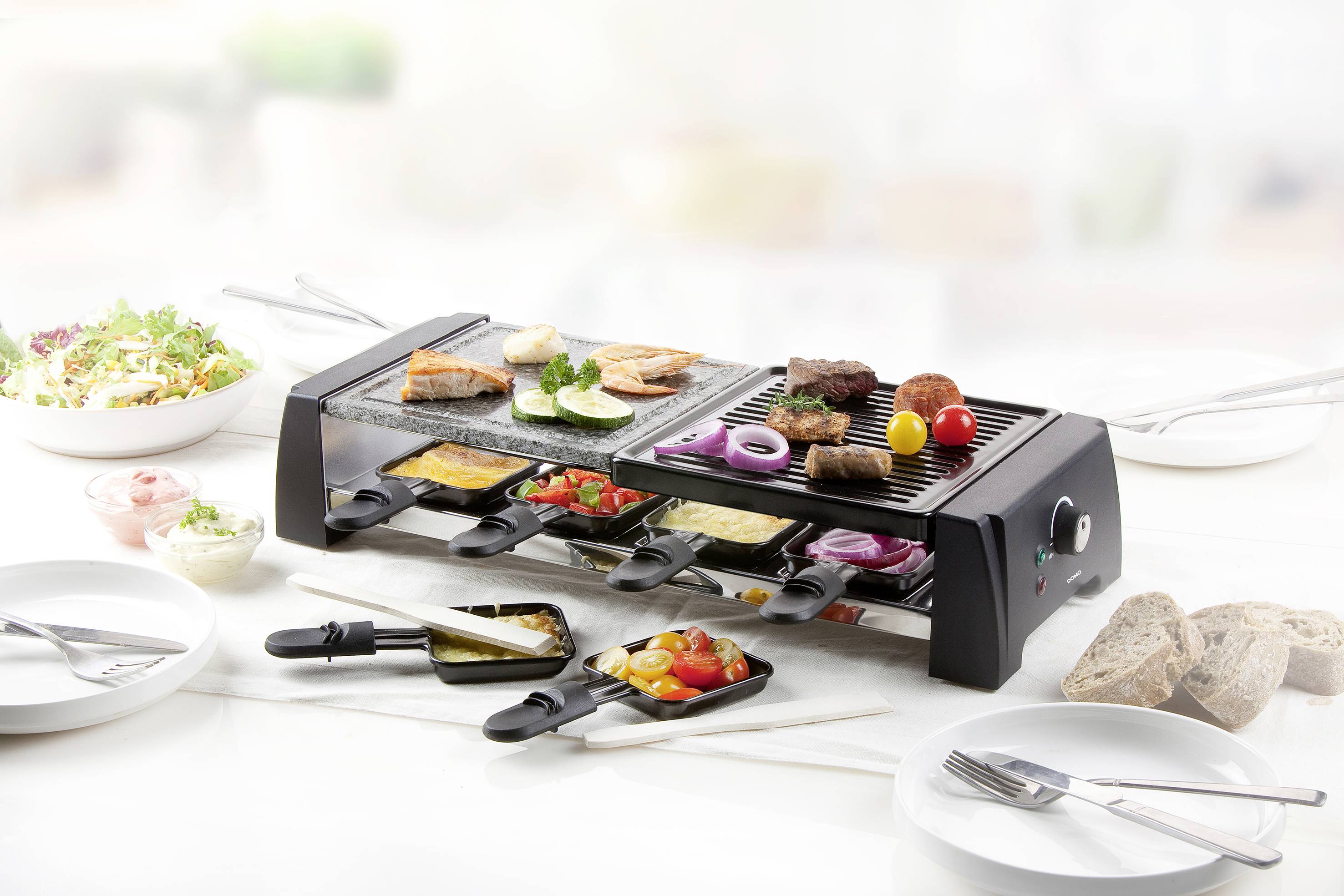 Een raclette-grill met verschillende ingrediënten zoals vlees, vis, groenten en kaas, omringd door bijgerechten zoals salade en brood.