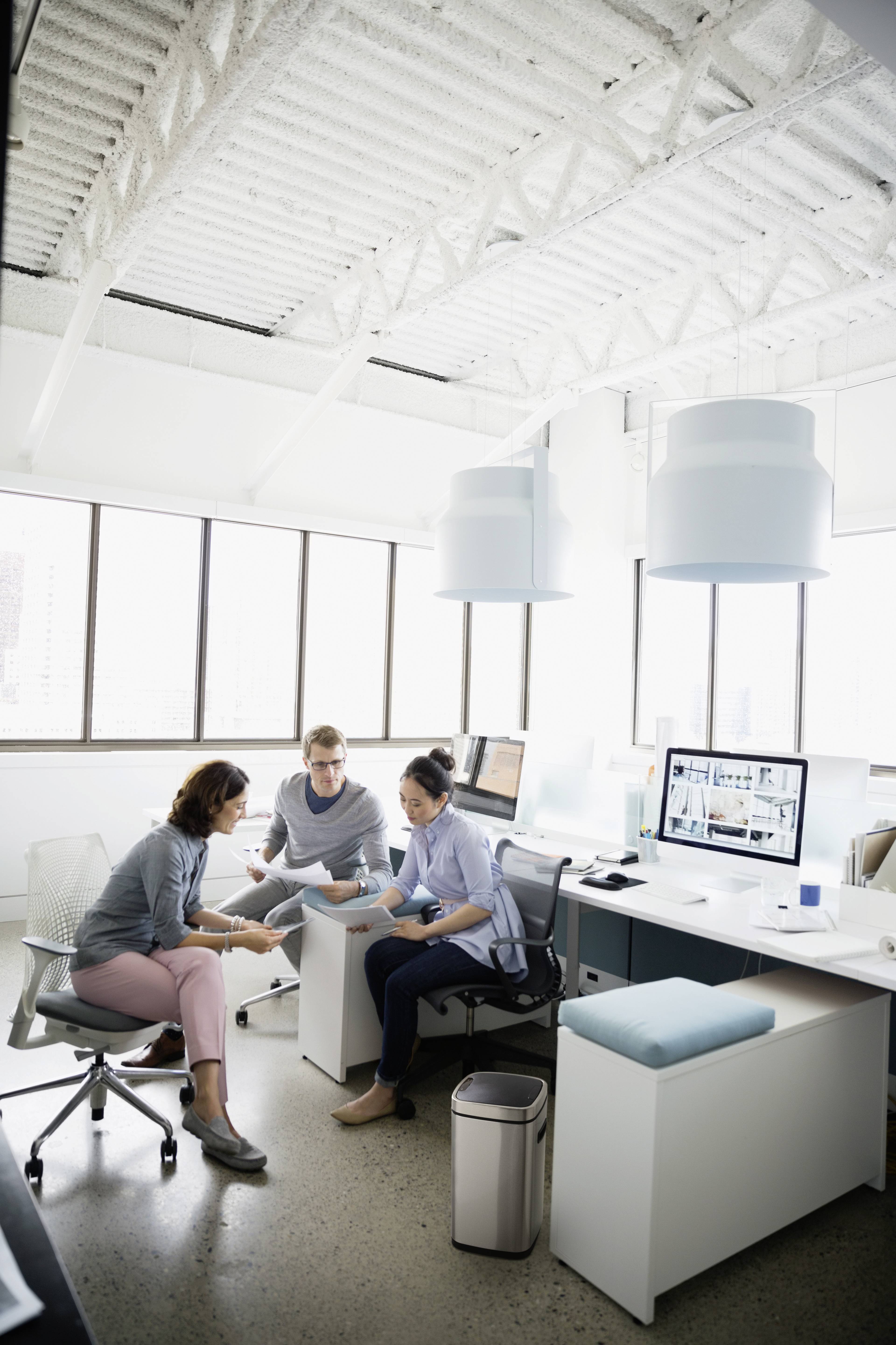 Drie personen werken samen in een modern kantoor. Ze zitten aan bureaus met computers, omringd door grote ramen.
