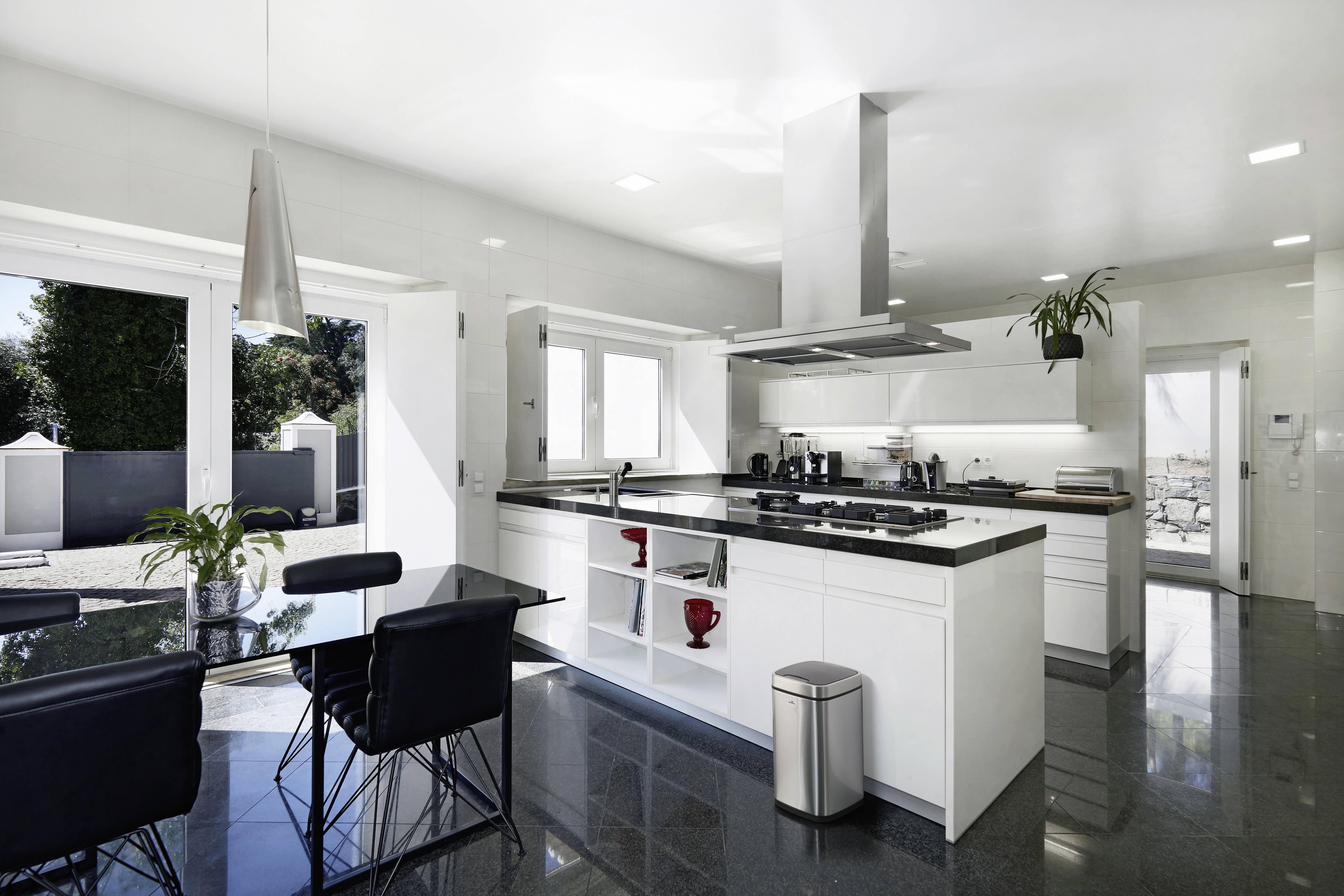 Moderne keuken met witte inrichting, groot raam en eettafel. Lichte, open indeling met planten en RVS-accenten.