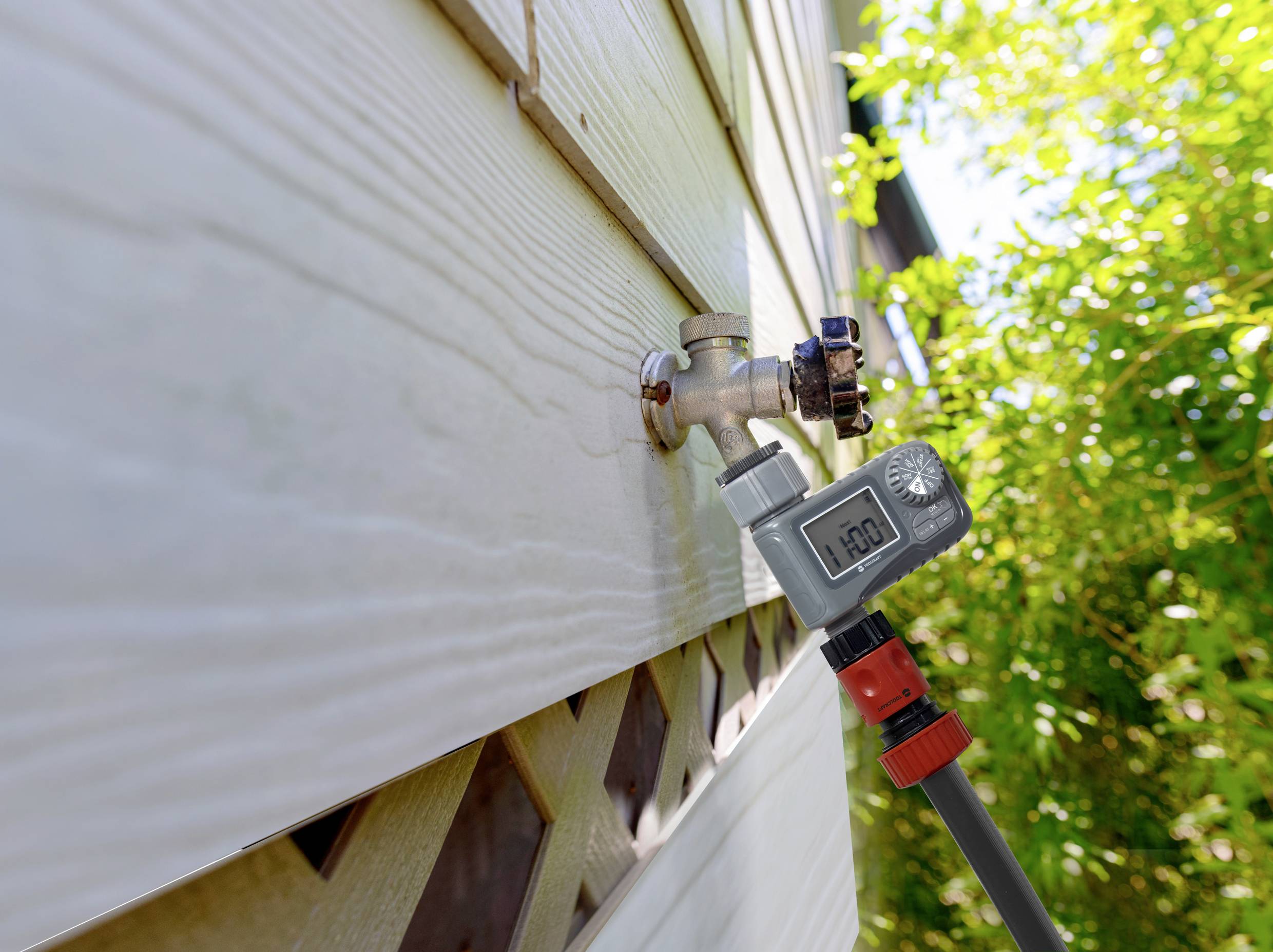Een digitale watermeter is bevestigd aan een buitenkraan. Het display toont '1.00'. Op de achtergrond zijn planten zichtbaar.