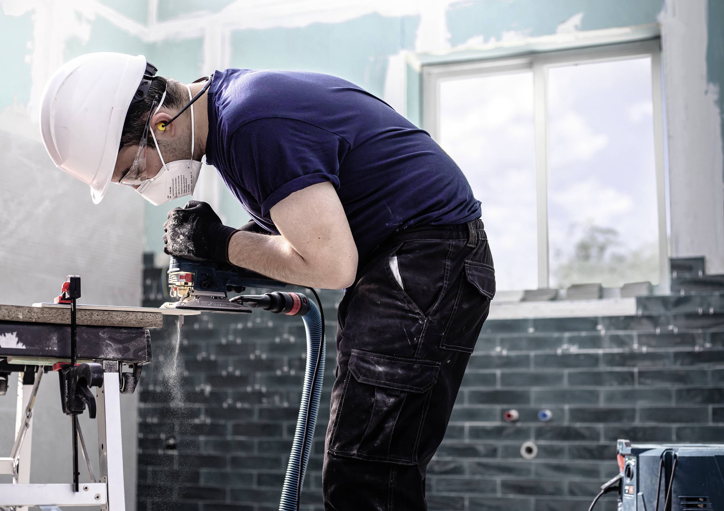 Een arbeider met helm en masker gebruikt een elektrisch gereedschap om hout te zagen in een halfgerenoveerde ruimte.