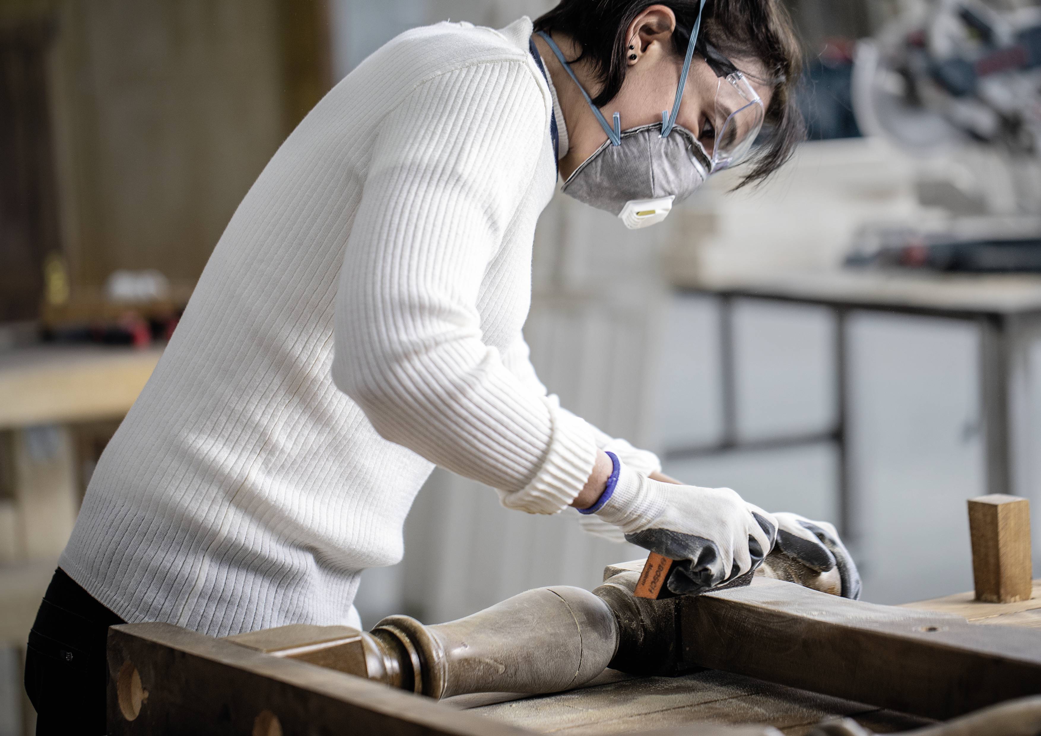 Een persoon in een witte trui slijpt een stuk hout in een werkplaats. Ze draagt een veiligheidsbril en een masker.