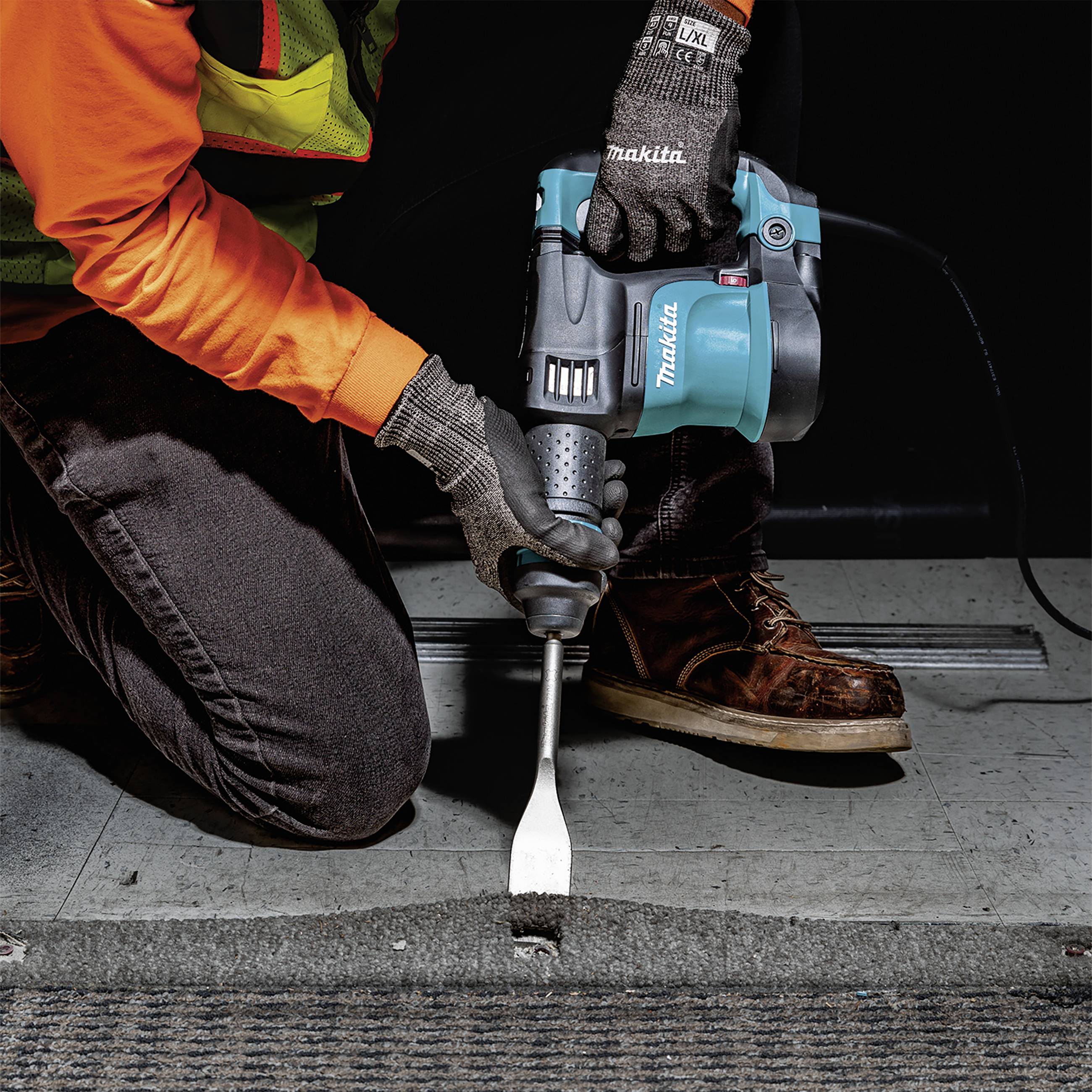 Een persoon verwijdert vloerbedekking met een elektrisch gereedschap in een industriële omgeving. Veiligheidsvest en -handschoenen worden gedragen.