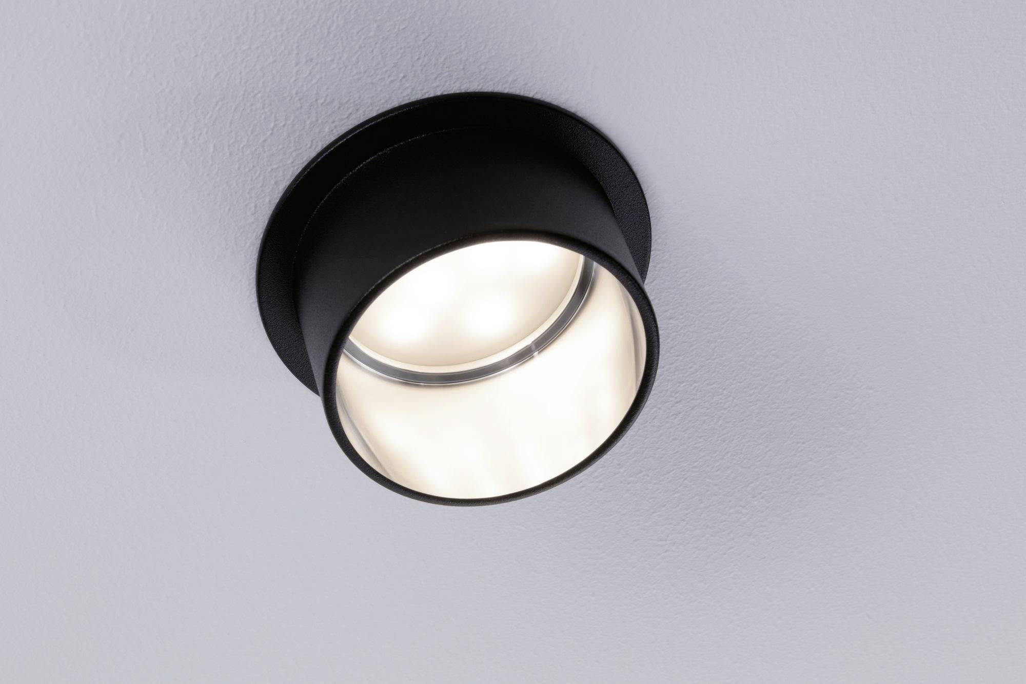 Een eenvoudige, zwarte plafondlamp is ingeschakeld en werpt warm licht op een witte plafond.