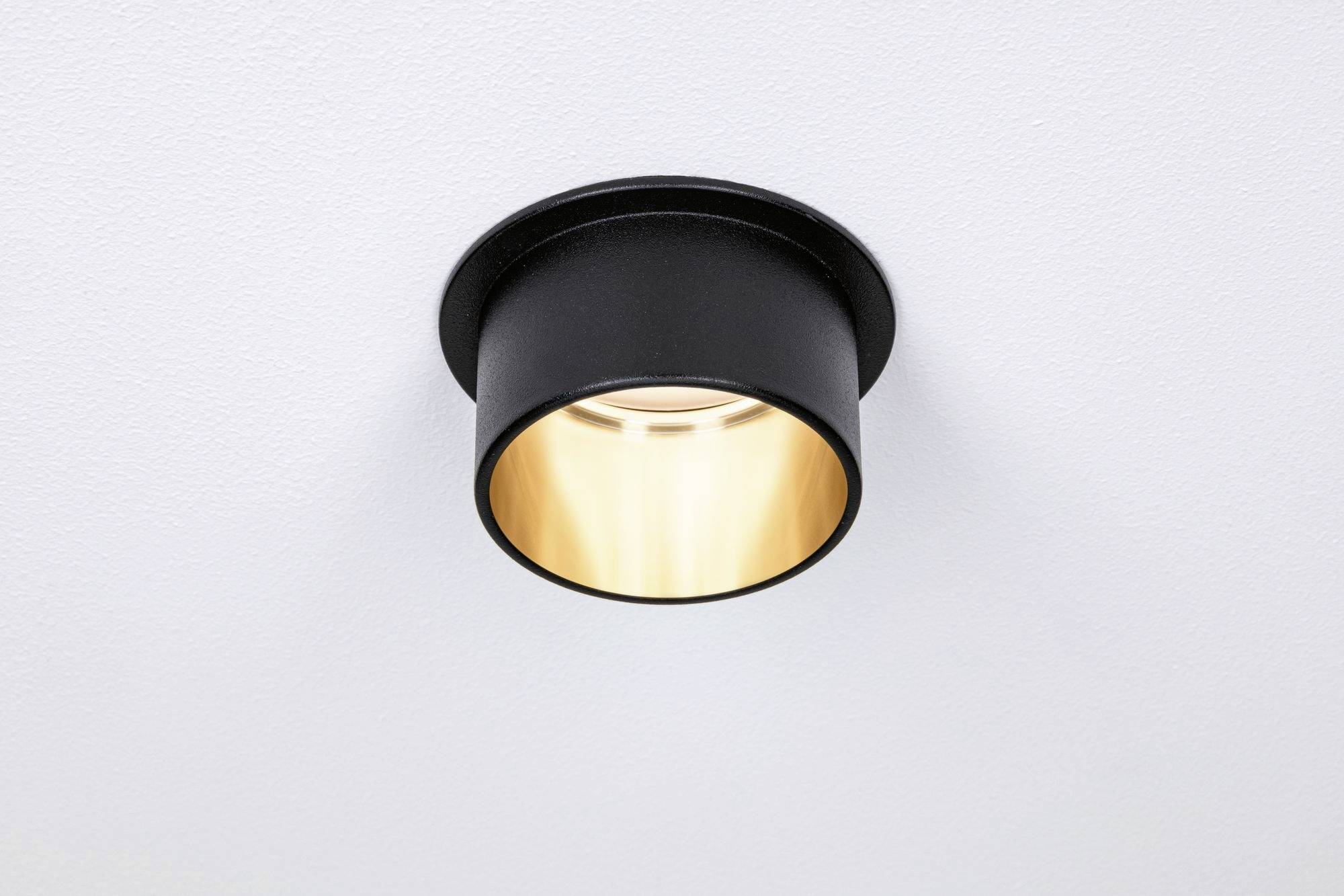 Een ronde, zwarte plafondlamp met warm licht, gemonteerd aan een witte plafond. Hij schijnt direct naar beneden.