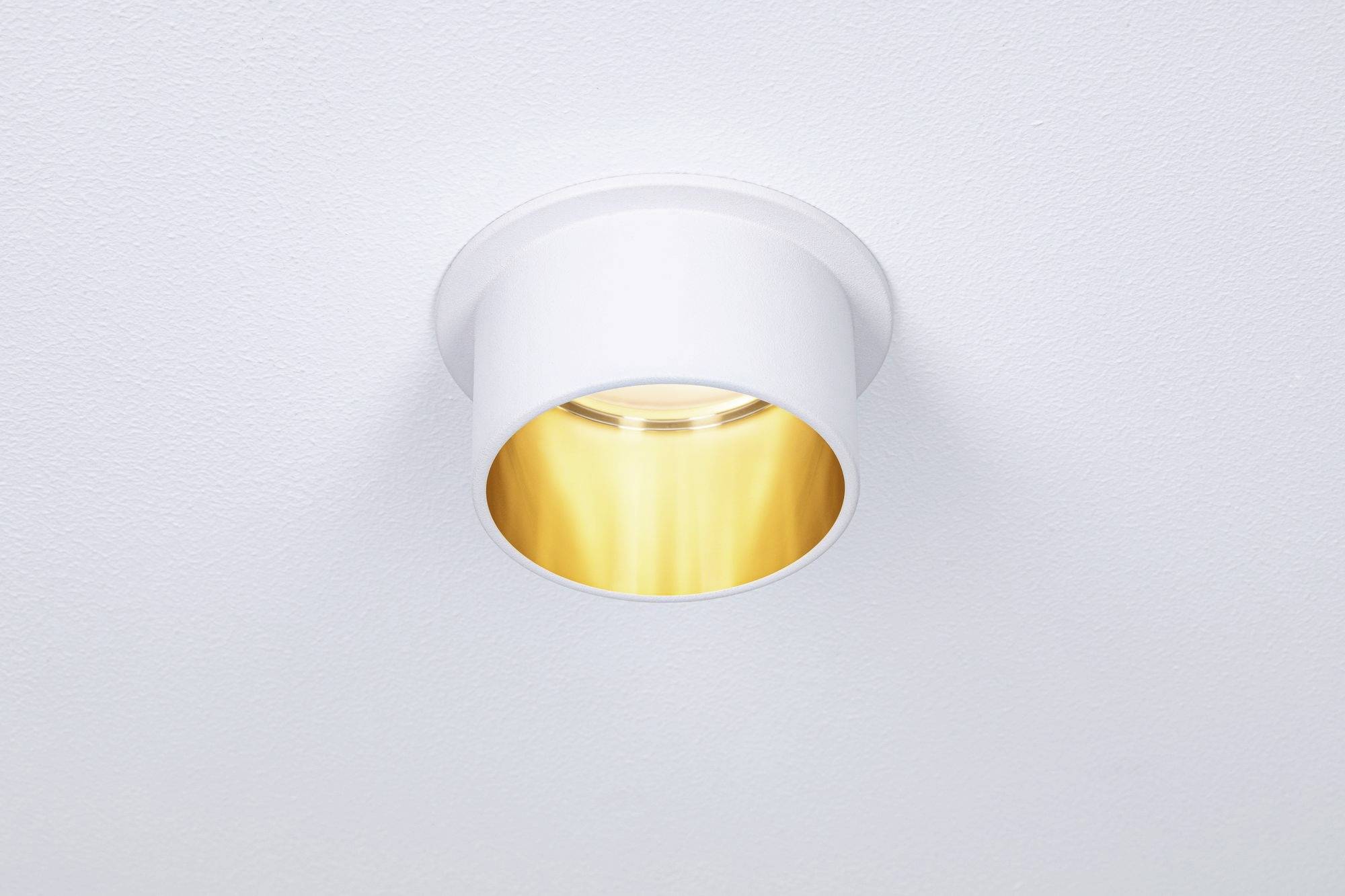 Een ronde, witte plafondlamp met een gouden binnenreflector die zacht, warm licht op een wit plafond projecteert.
