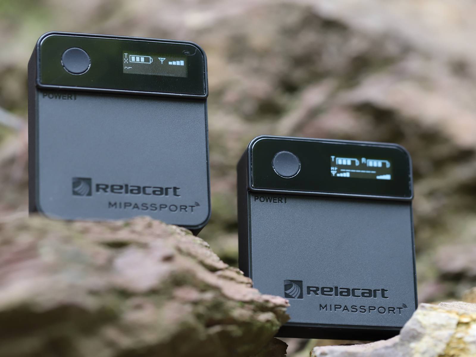 Twee draagbare audio-apparaten met display en het logo 'Relacart MIPASSPORT' op rotsen. Ze zijn ingeschakeld en tonen het batterijniveau.