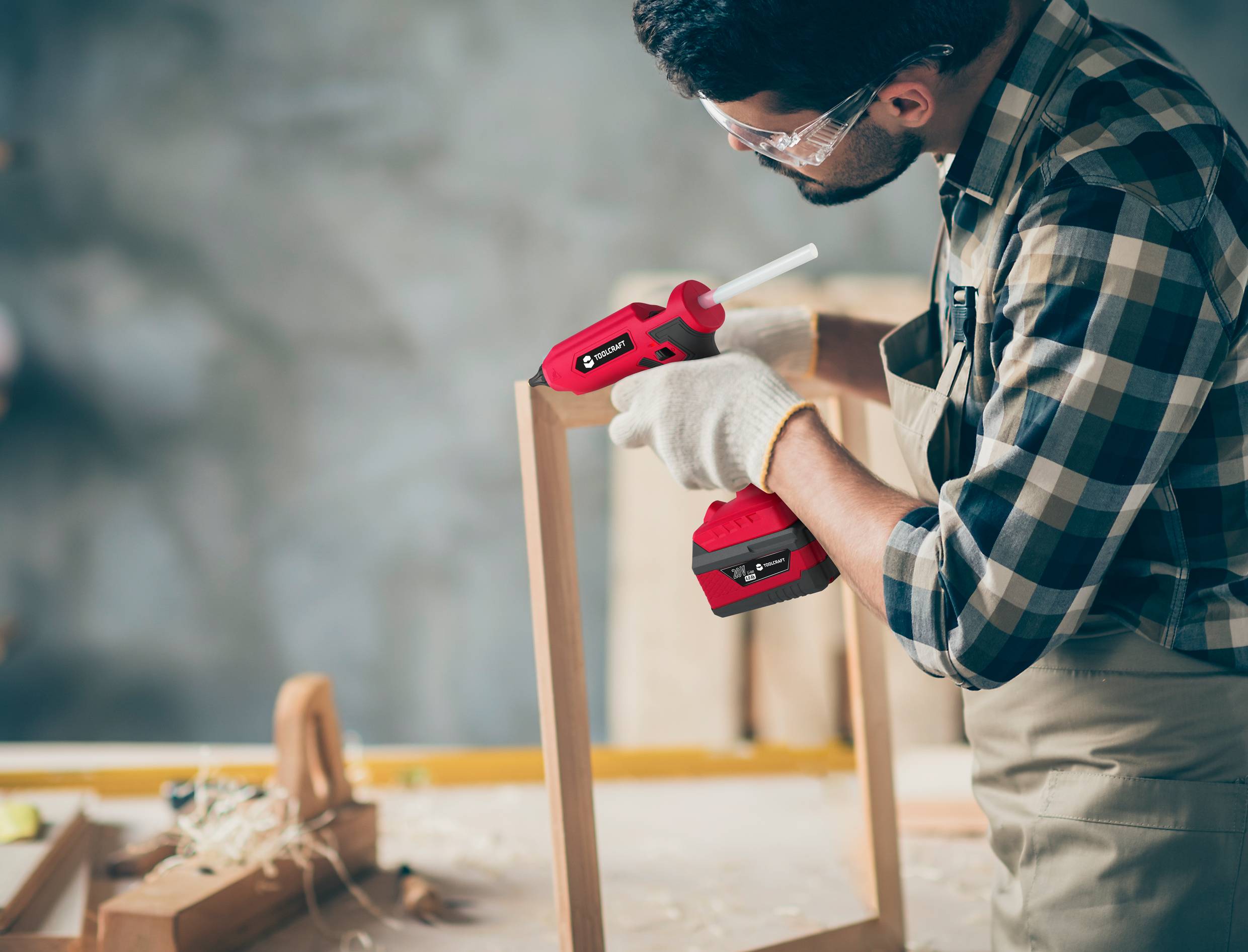Een man met veiligheidsbril en handschoenen gebruikt een rode lijmpistool om een houten frameonderdeel aan elkaar te bevestigen.