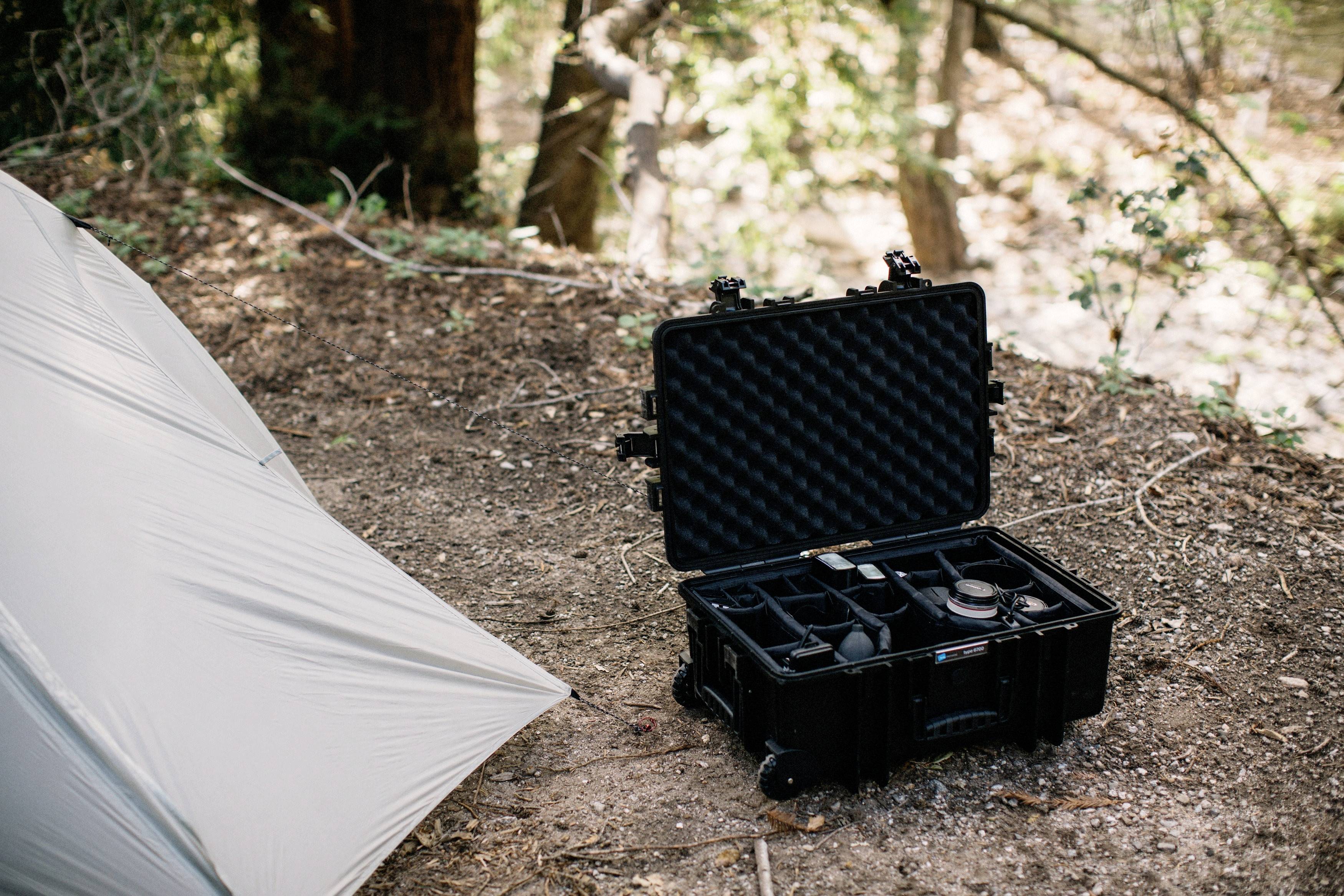 Een open zwarte koffer met cameratoebehorenb op de bosbodem, naast een tent in een bebost gebied.