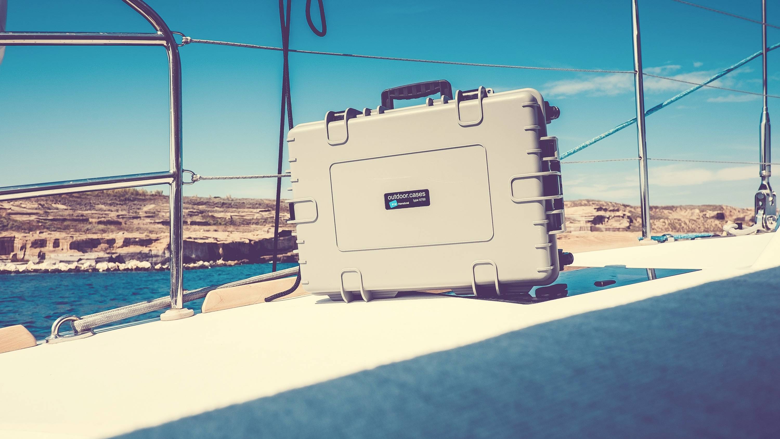 Een zilveren koffer ligt op het dek van een zeilboot die vaart op een kalme zee. Op de achtergrond is de kust zichtbaar.