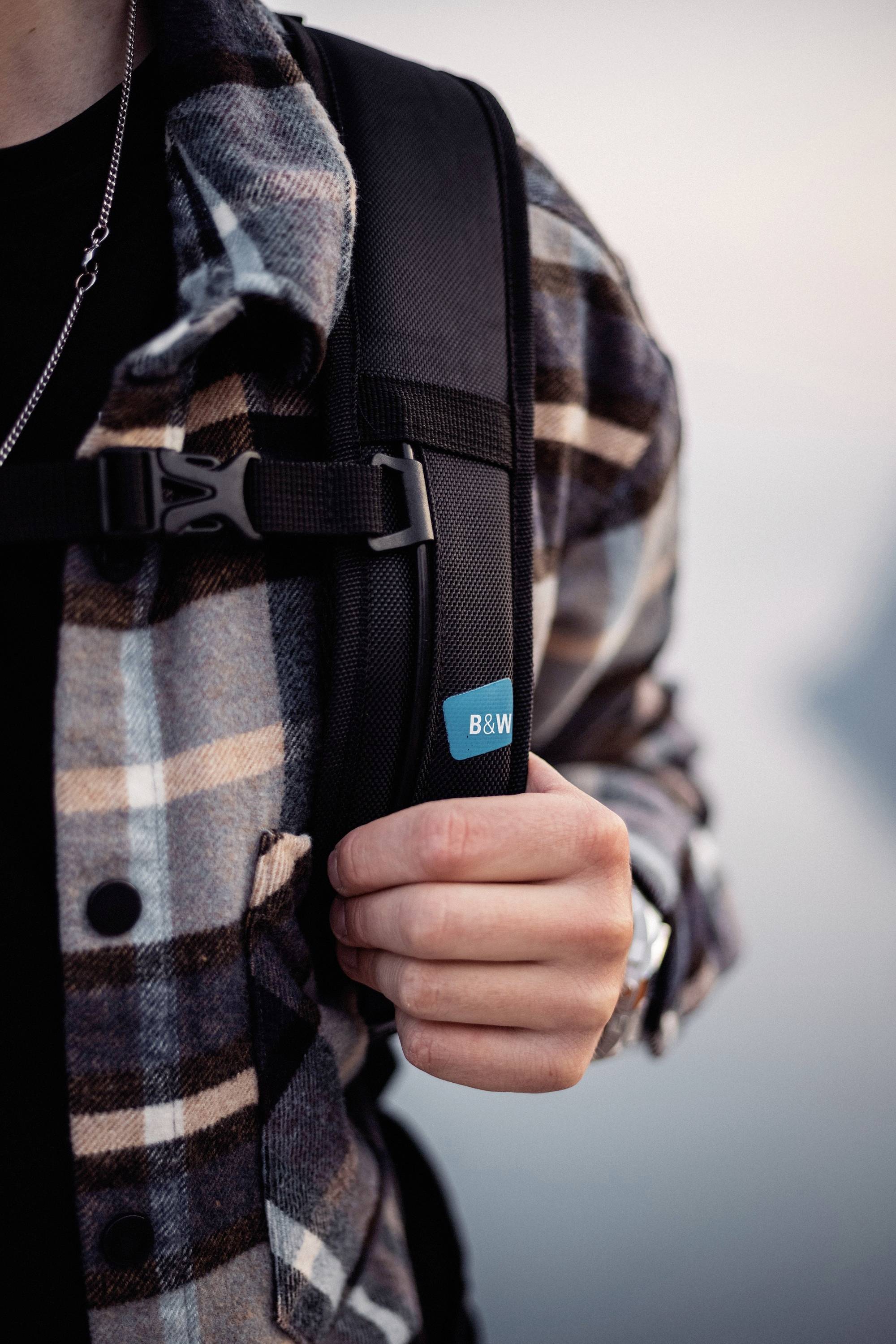 Persoon draagt geruit overhemd en zwarte rugzak, houdt de drager vast met zijn rechterhand. Sfeervolle scène met een onscherpe achtergrond.