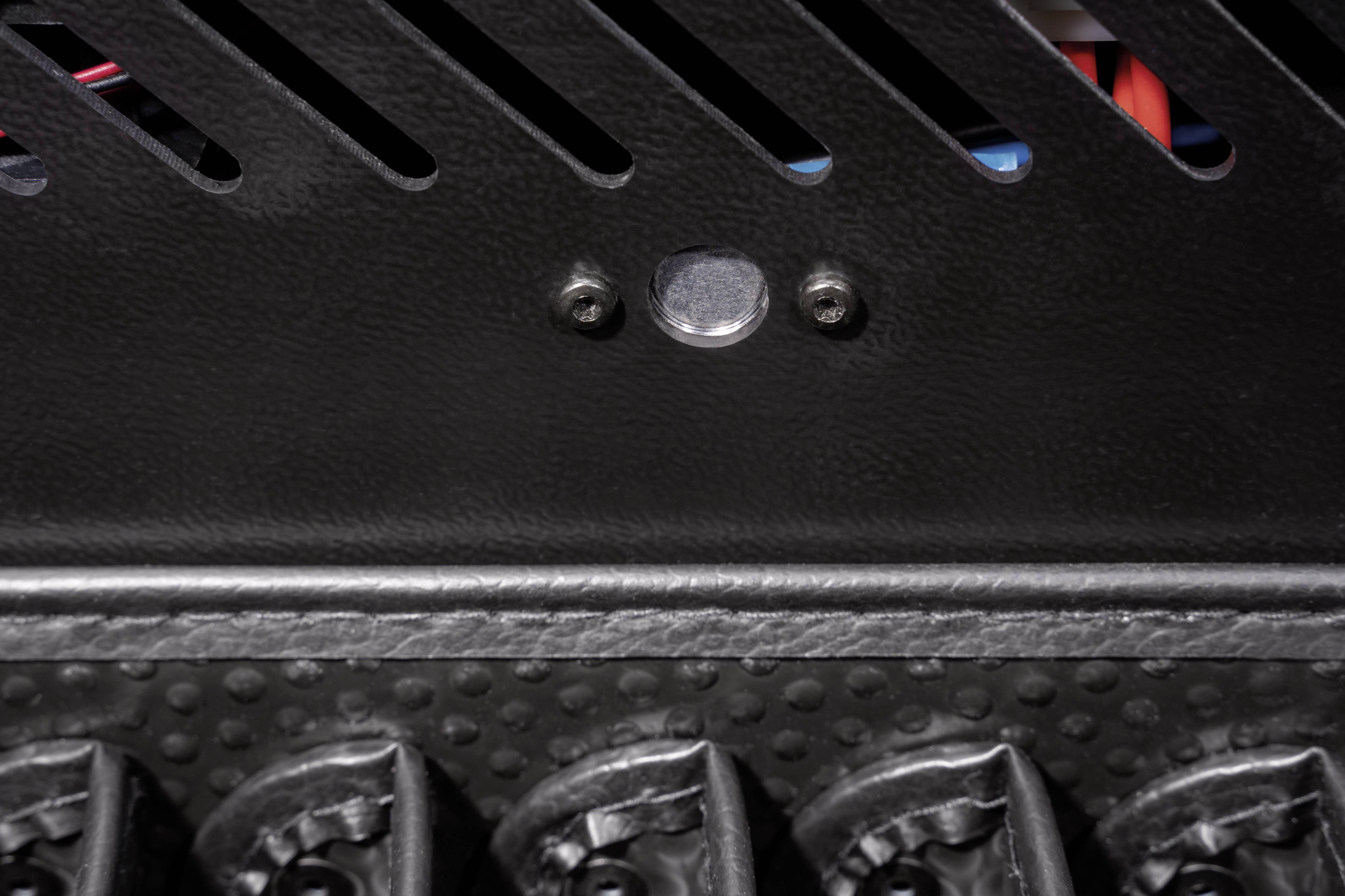 Close-up van een zwarte metalen plaat met geribbelde lijnen en een ronde metalen knop. Daaronder zijn gestructureerde verhogingen zichtbaar.