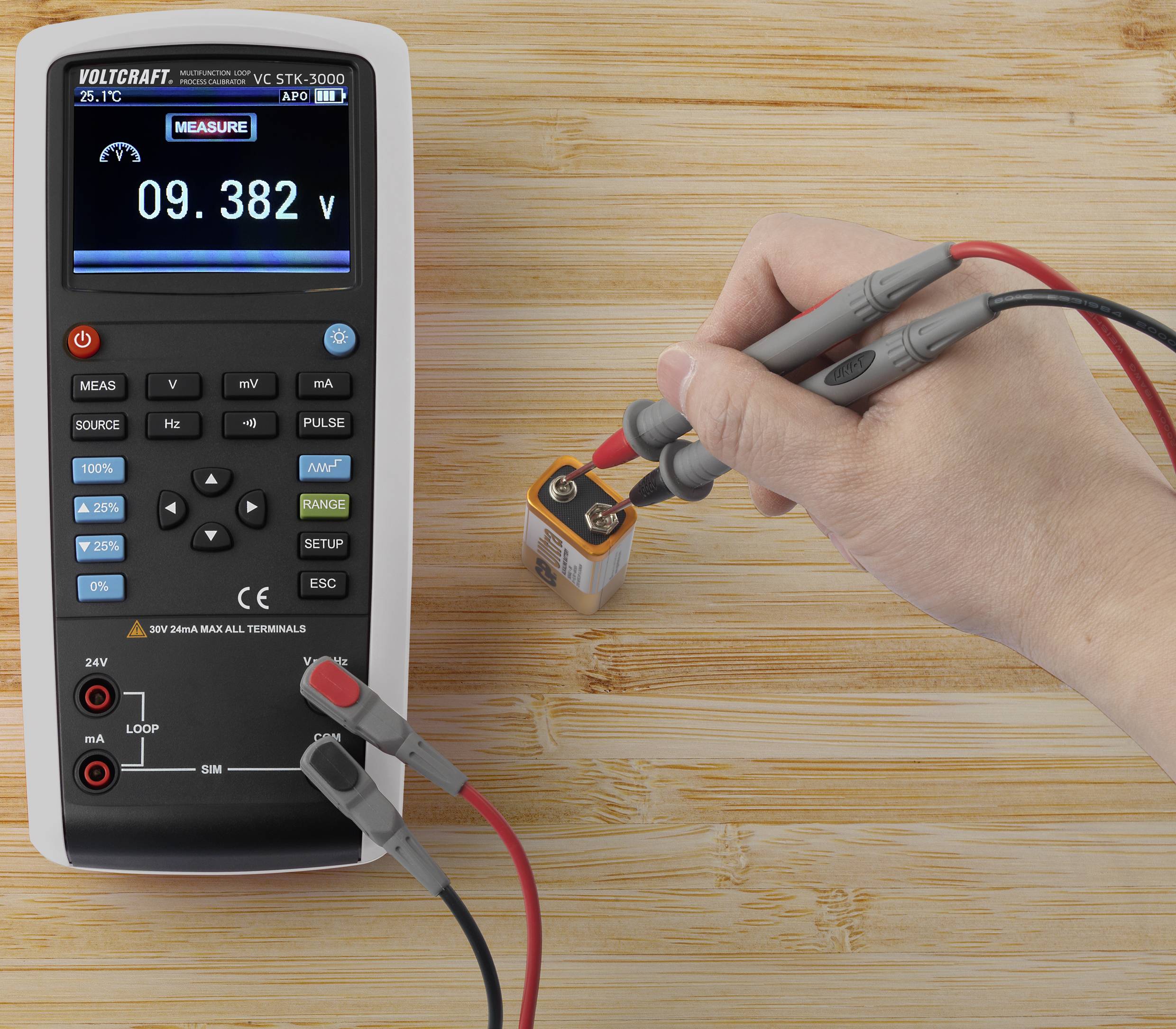 Een multimeter toont '09,382 V' op het display. Een hand meet met meetpennen de spanning van een 9-volt batterij.