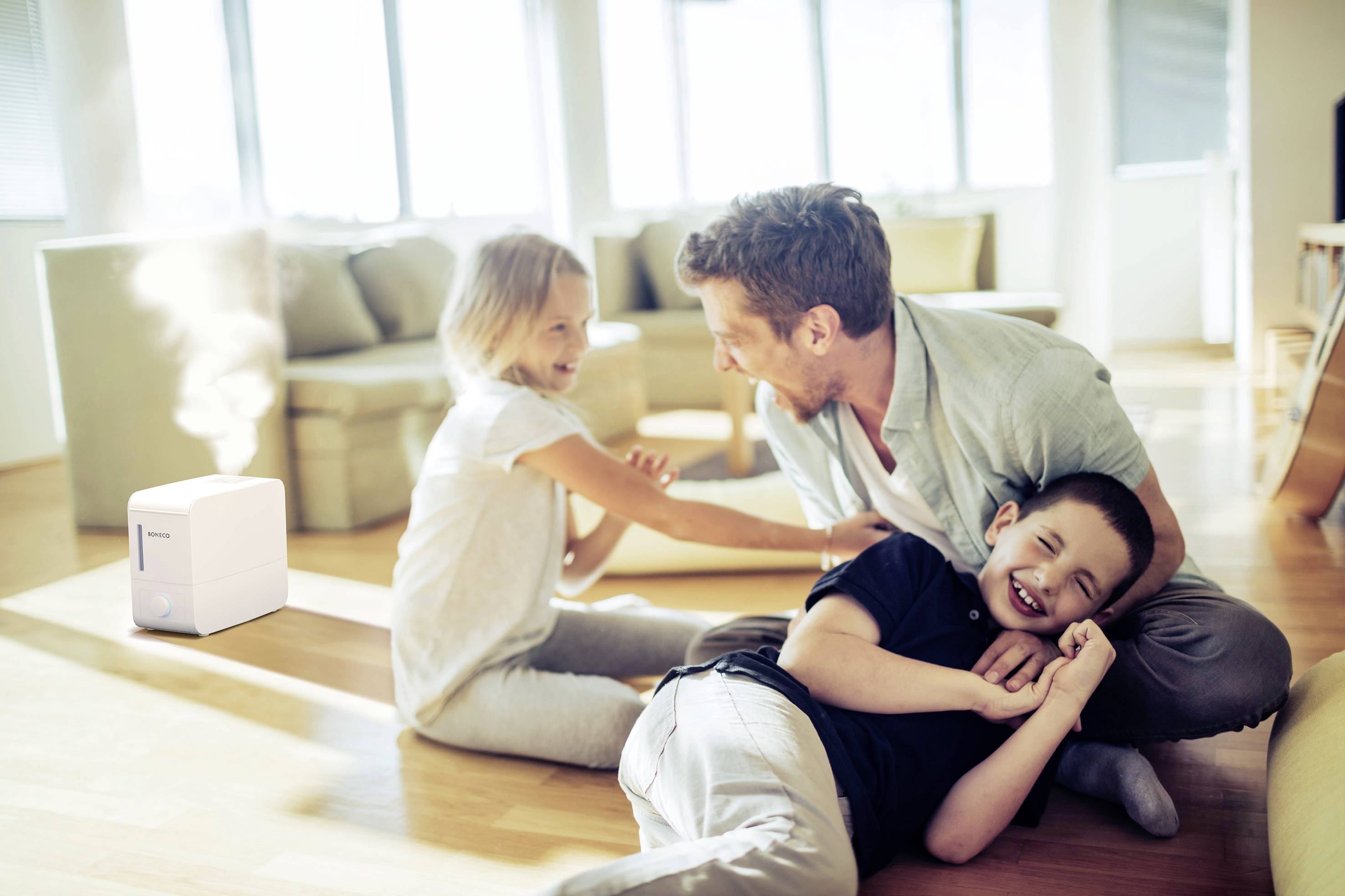 Een man en twee kinderen lachen en spelen op de woonkamervloer, terwijl op de achtergrond een luchtbevochtiger stoom uitblaast.