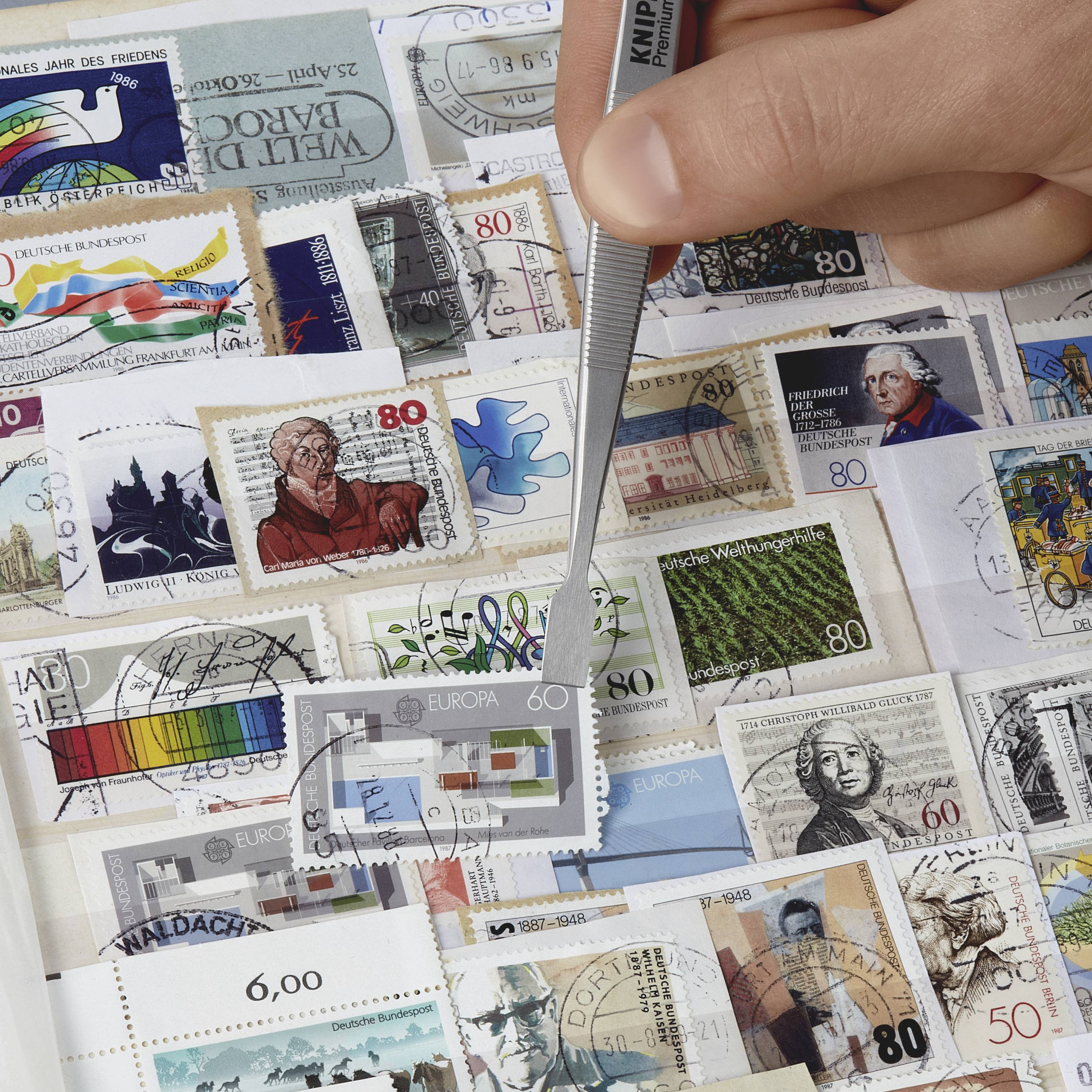 Een persoon gebruikt een pincet om een postzegel te nemen uit een verscheidenheid aan kleurrijke en gestempelde postzegelontwerpen.