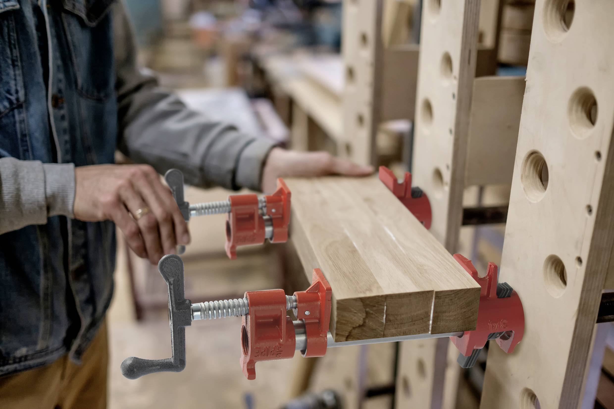 Een persoon bevestigt een plank met rode schroefklemmen aan een verticaal frame. Op de achtergrond is een werkplaats zichtbaar.