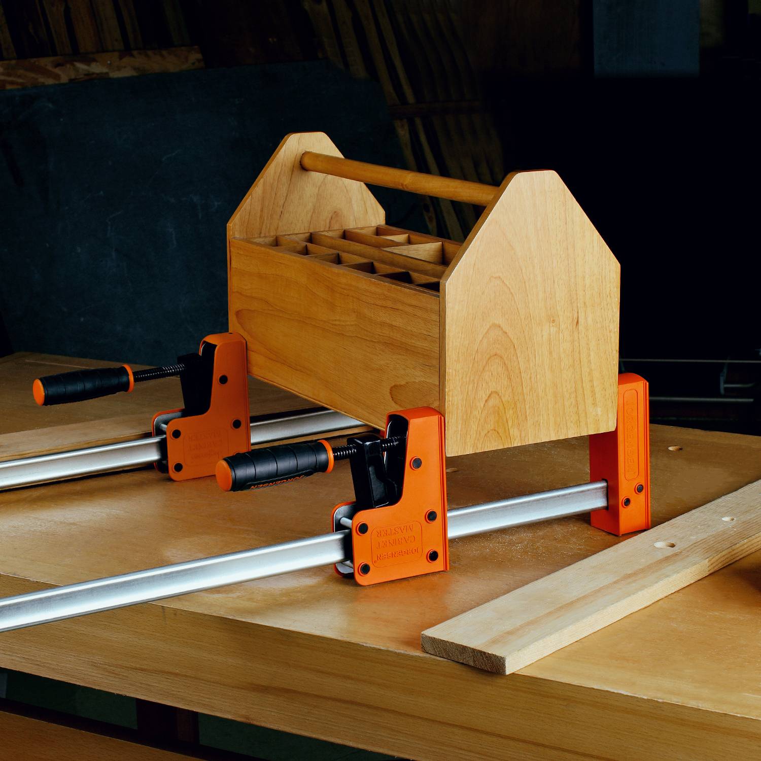 Kleine houten gereedschapskist met handgreep wordt met klemmen vastgemaakt op een werkbank. Op de achtergrond zijn houten platen zichtbaar.