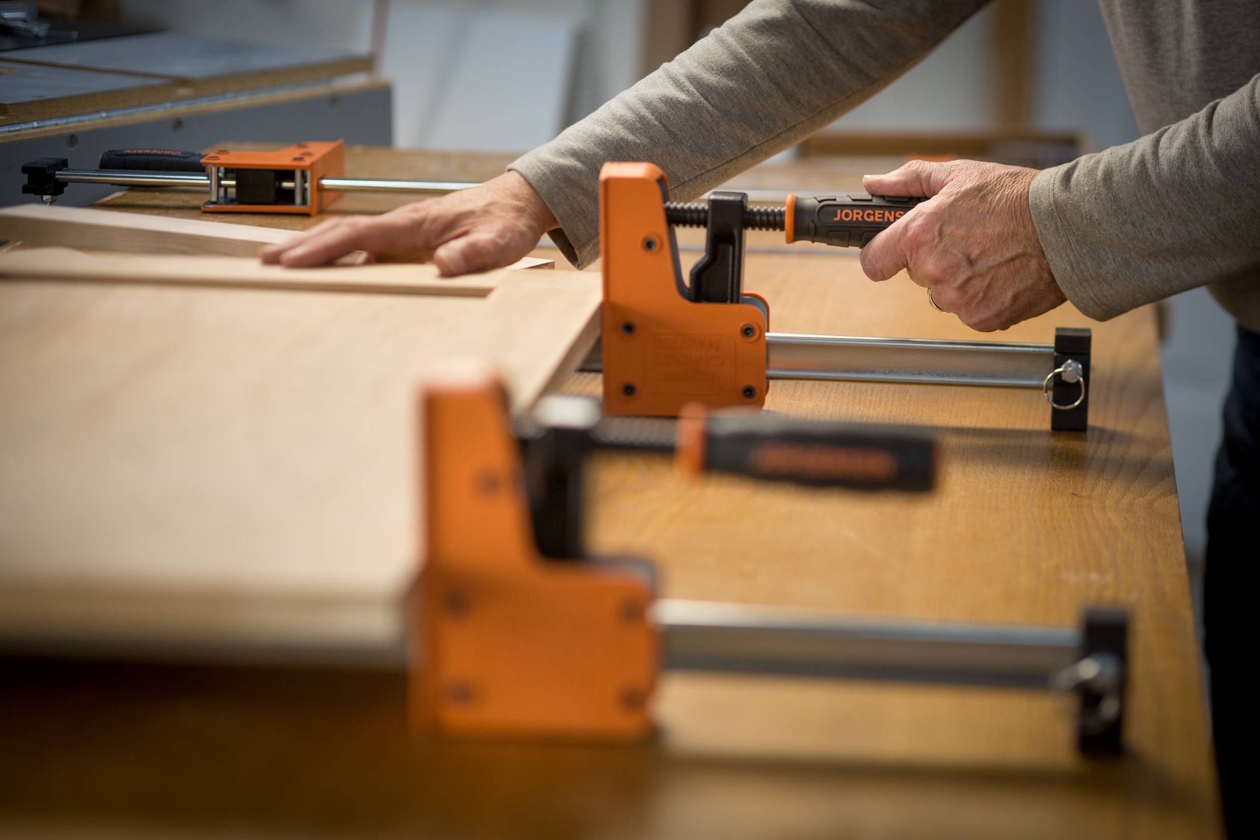 Een persoon gebruikt oranje klemmen om een houten plank op een tafel vast te zetten. Handen stellen de klemmen bij.