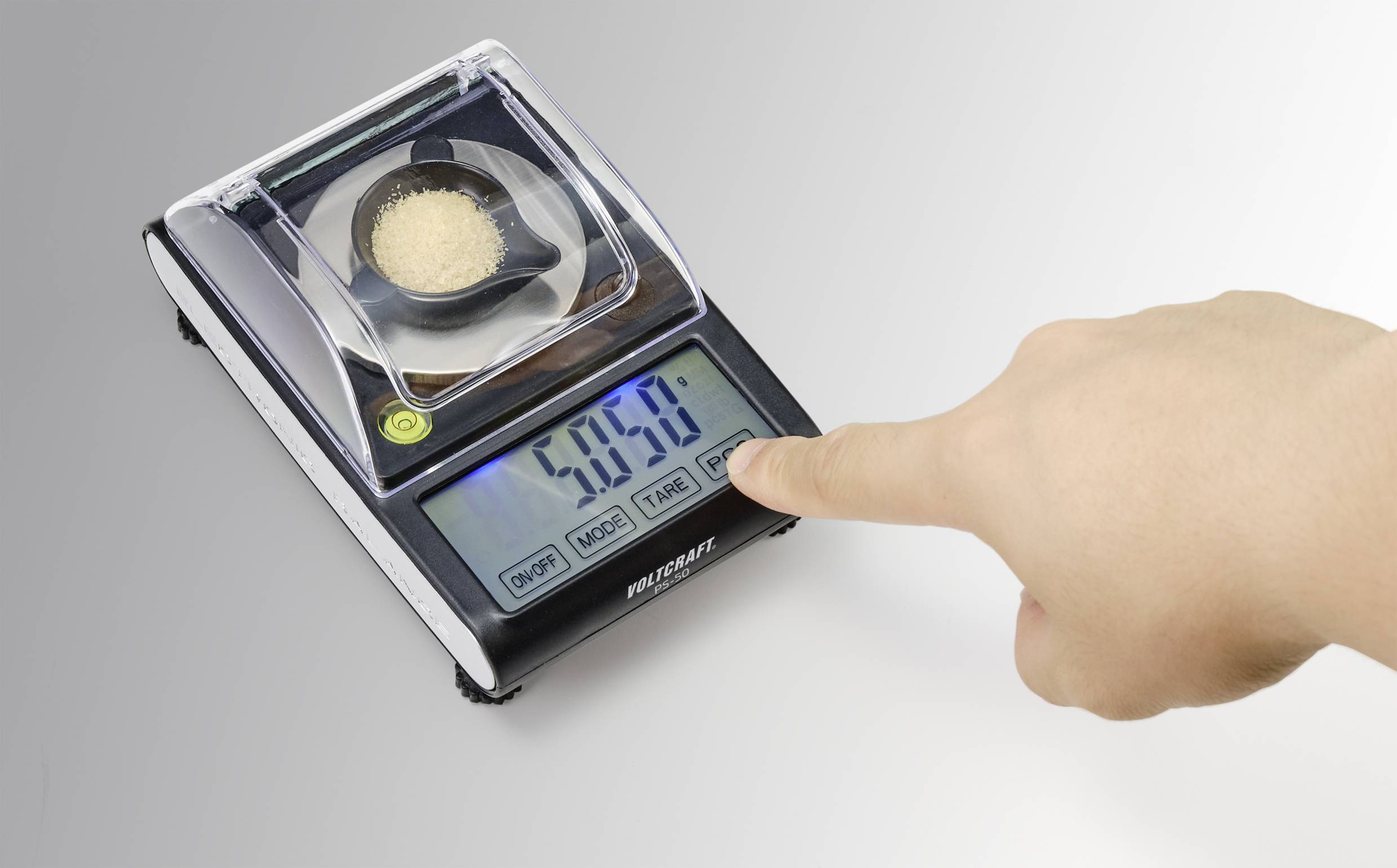 Een digitale weegschaal toont een gewicht van 5,859 gram. Een hand drukt op de 'TARE'-toets. Op de weegschaal staat een kleine container met poeder.