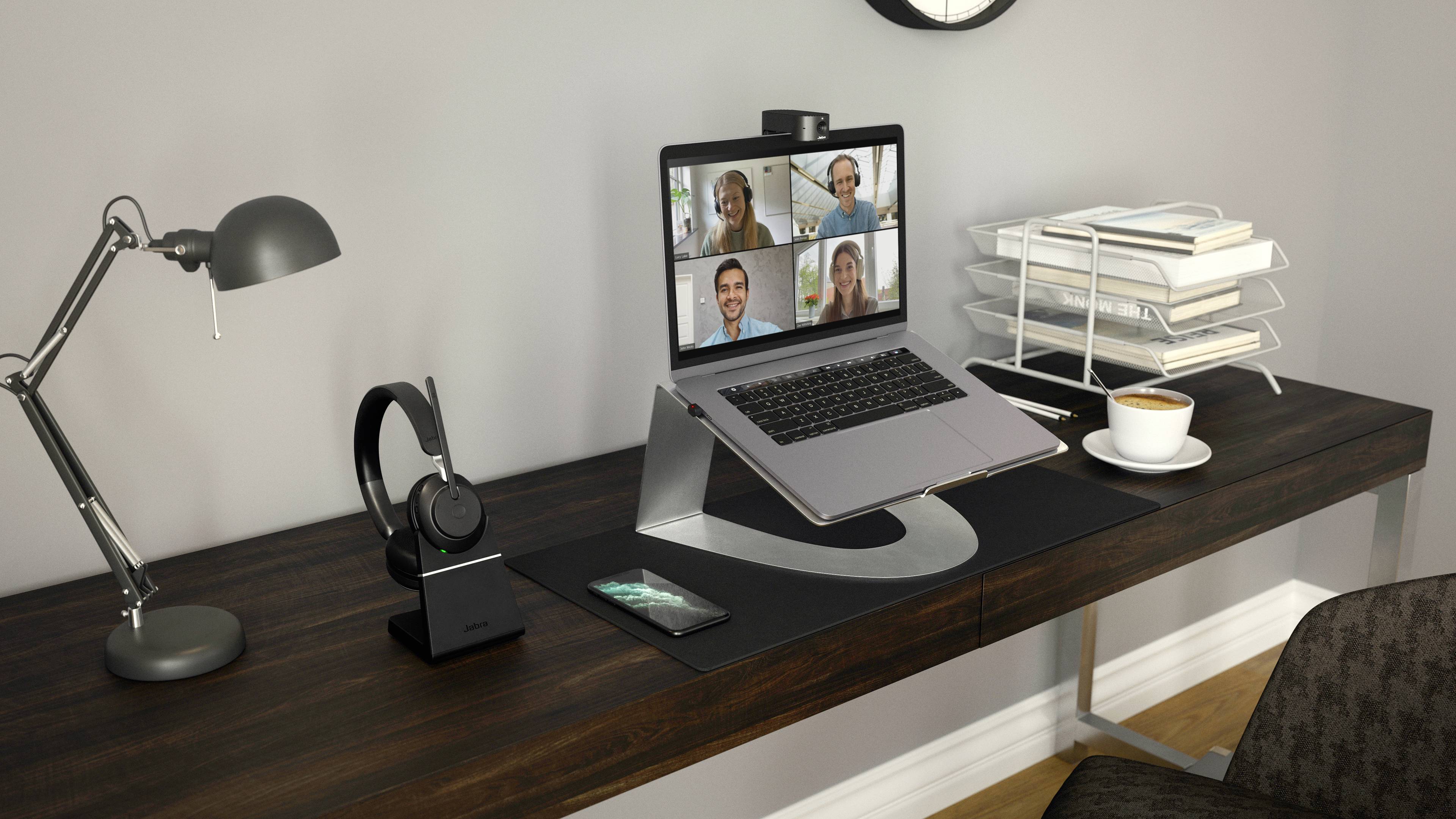 Een bureau met een laptop waarop een videogesprek met vier personen plaatsvindt. Ernaast staan een kopje, koptelefoon en een lamp.