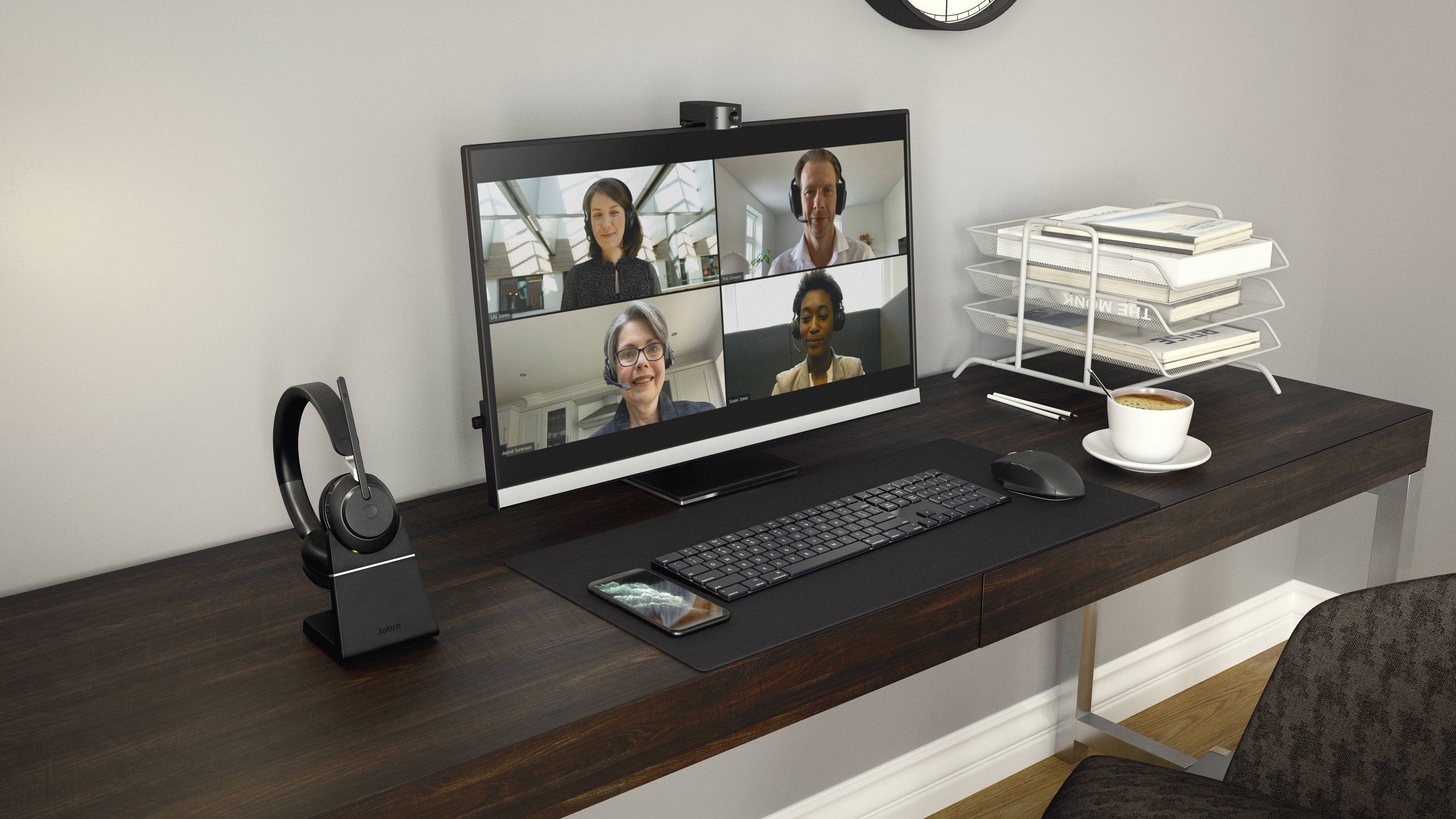 Een bureau met een computerscherm waarop een videoconferentie met vier personen wordt weergegeven. Ernaast liggen een headset en een kopje koffie.