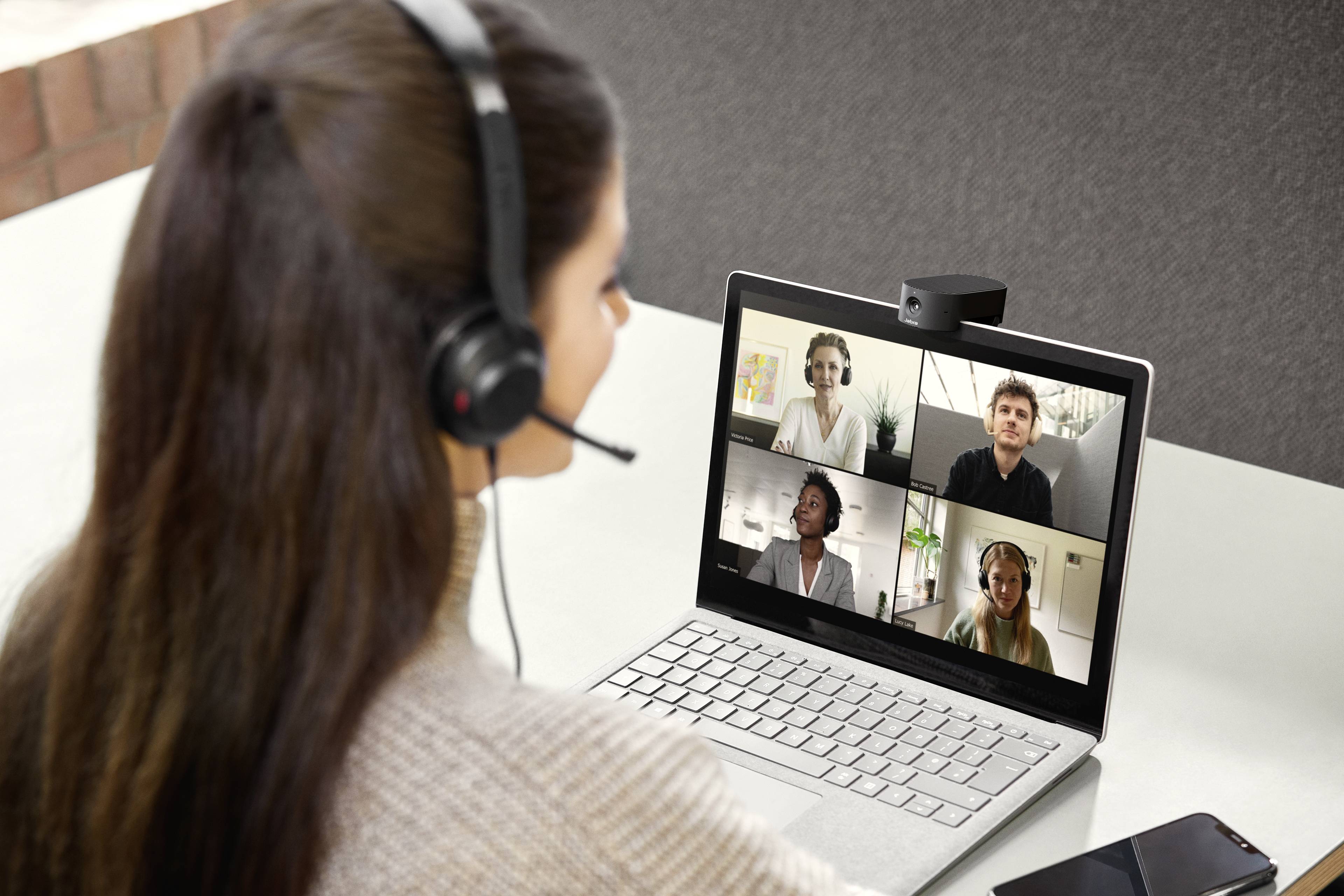 Een vrouw met een headset neemt deel aan een videovergadering op haar laptop. Vier personen zijn zichtbaar op het scherm.