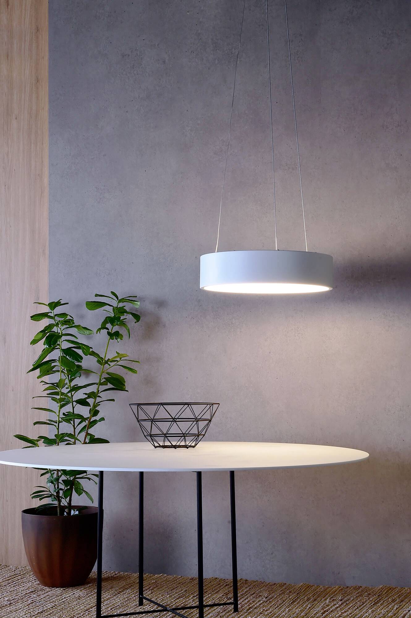 Een ronde tafel met een decoratieve, lege schaal erop. Een hanglamp schijnt van bovenaf. Op de achtergrond staat een plant.