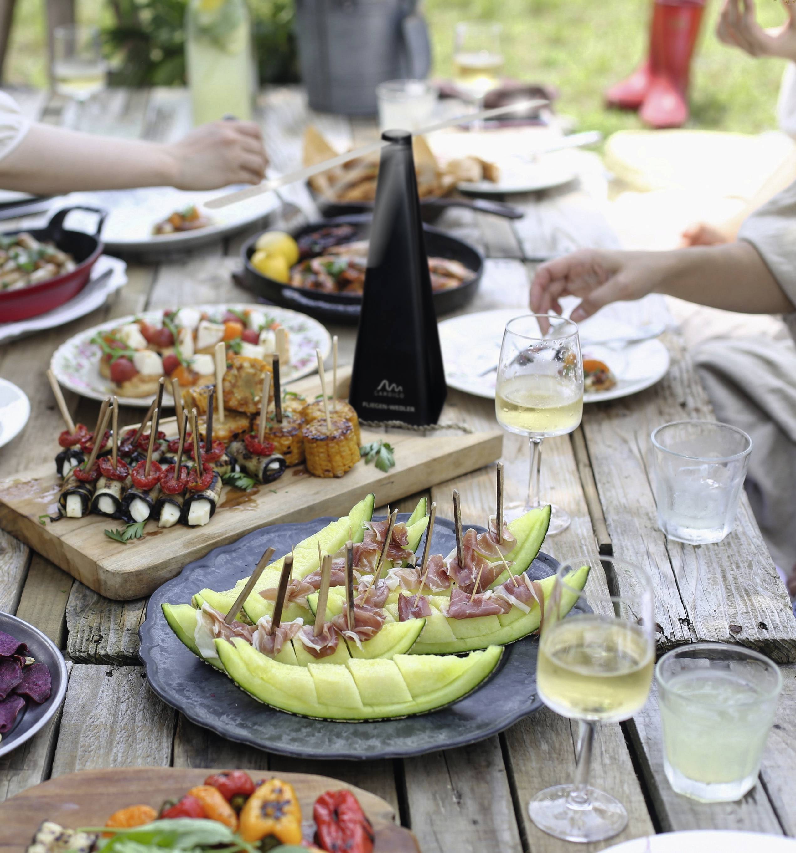 Een gedekte houten tafel buiten met verschillende gerechten: meloen met ham, gevulde paprika's, gegrilde groenten en glazen met dranken.