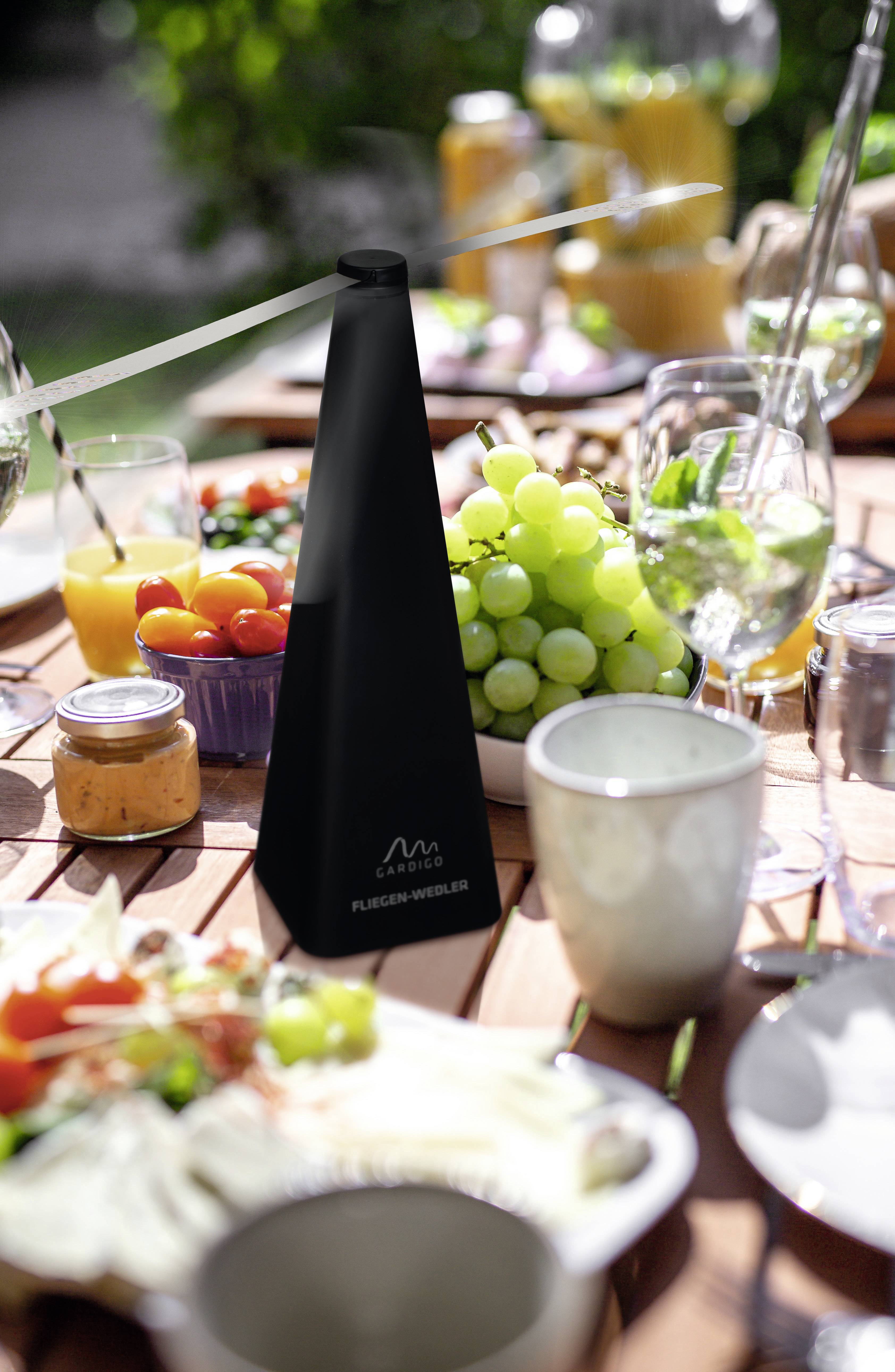 Een tafel buiten is gedekt met vers fruit, dranken en bestek. Op de voorgrond staat een zwart, modern keukenhulpmiddel.