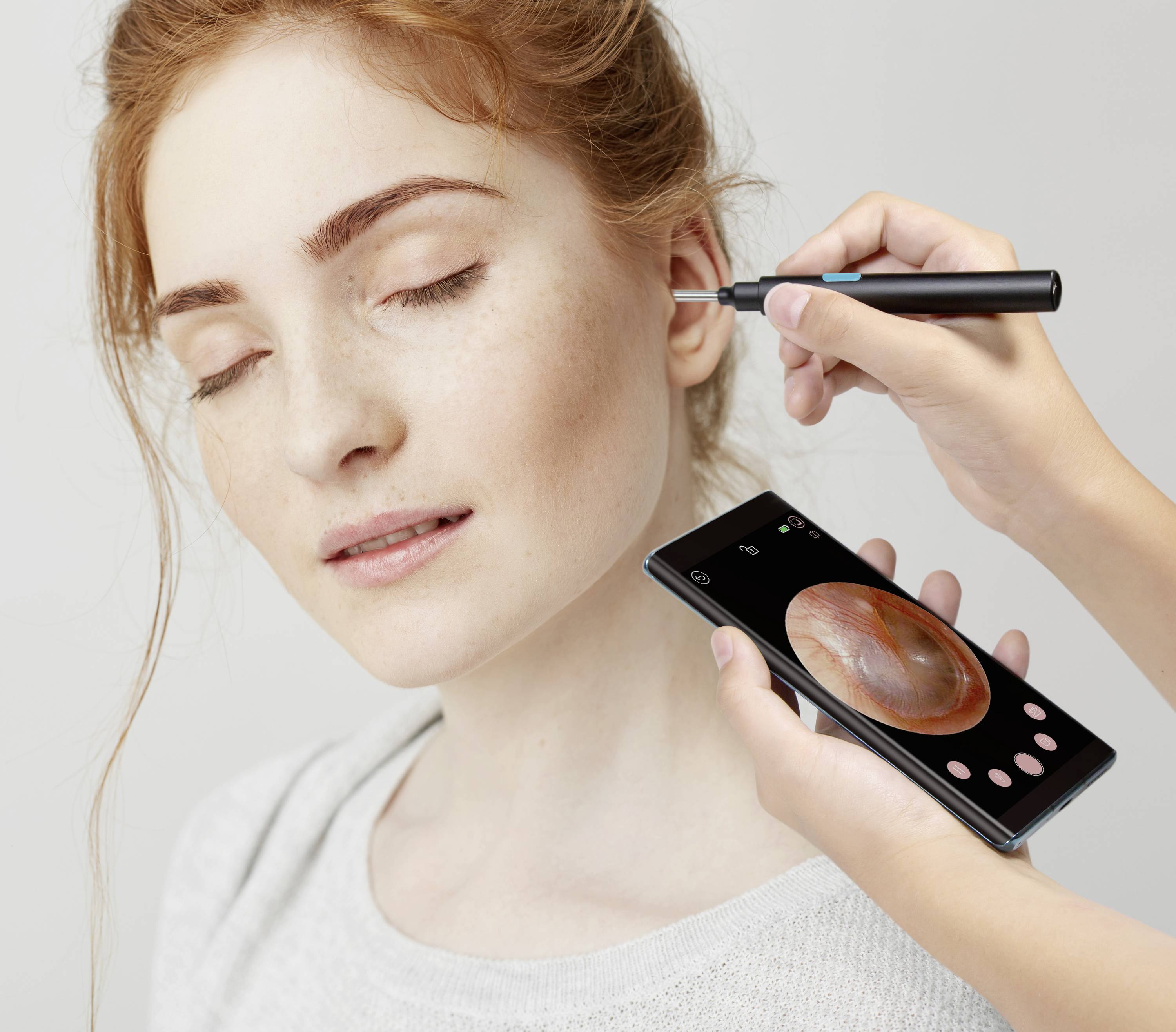 Een vrouw heeft haar ogen gesloten terwijl een hand een apparaat gebruikt om haar oren te reinigen. Een mobiele telefoon toont een close-up.