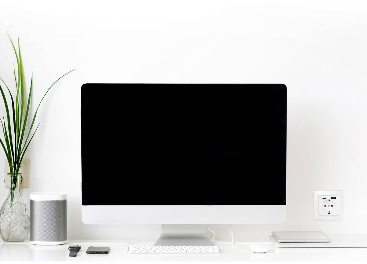Een moderne schrijftafel met een groot computerscherm. Ernaast staan een plant, een speaker en een laptop.