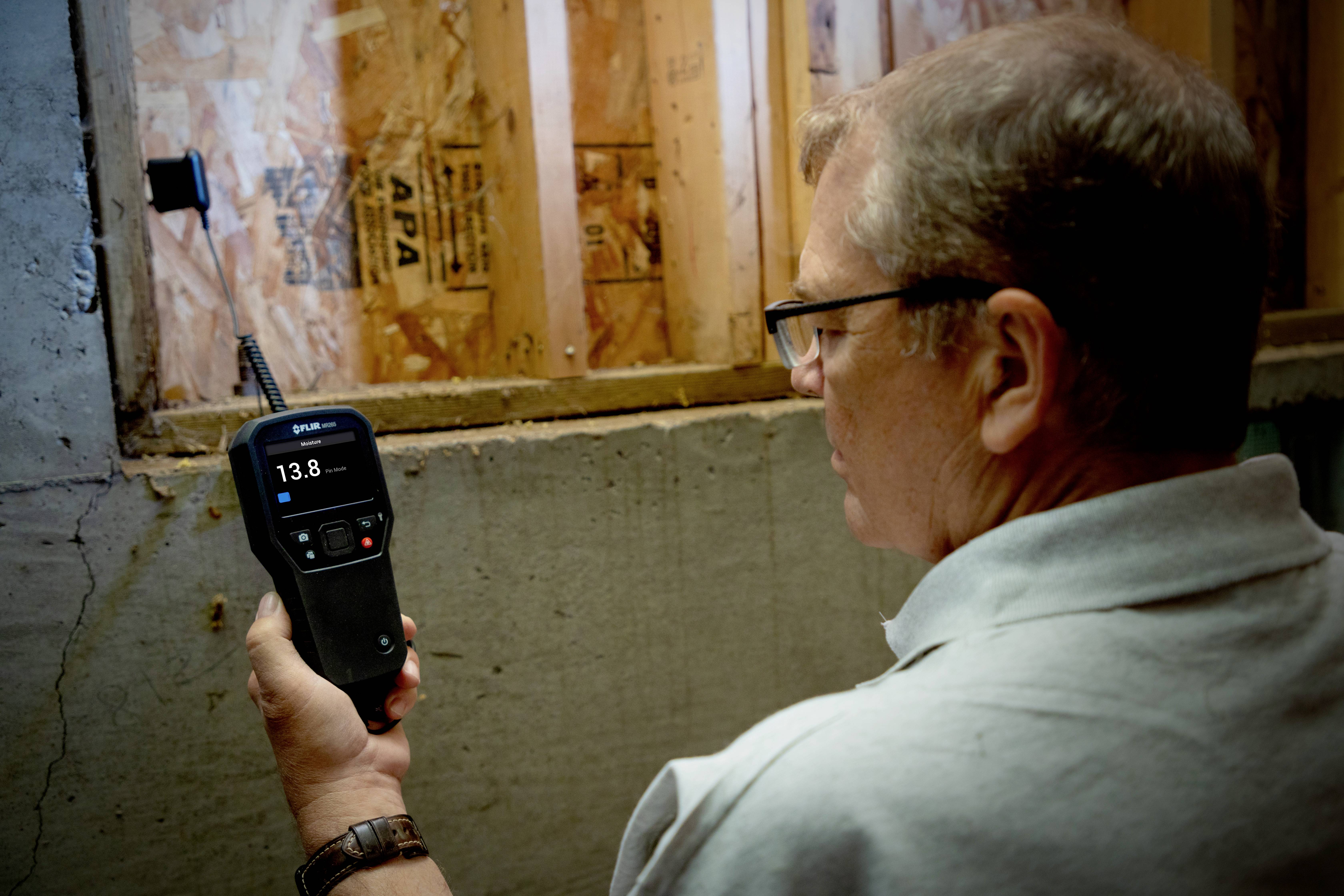 Een man houdt een vochtmeter tegen een muur in de kelder, die 13,8 aanwijst. De muur bestaat uit onbekleed hout en beton.