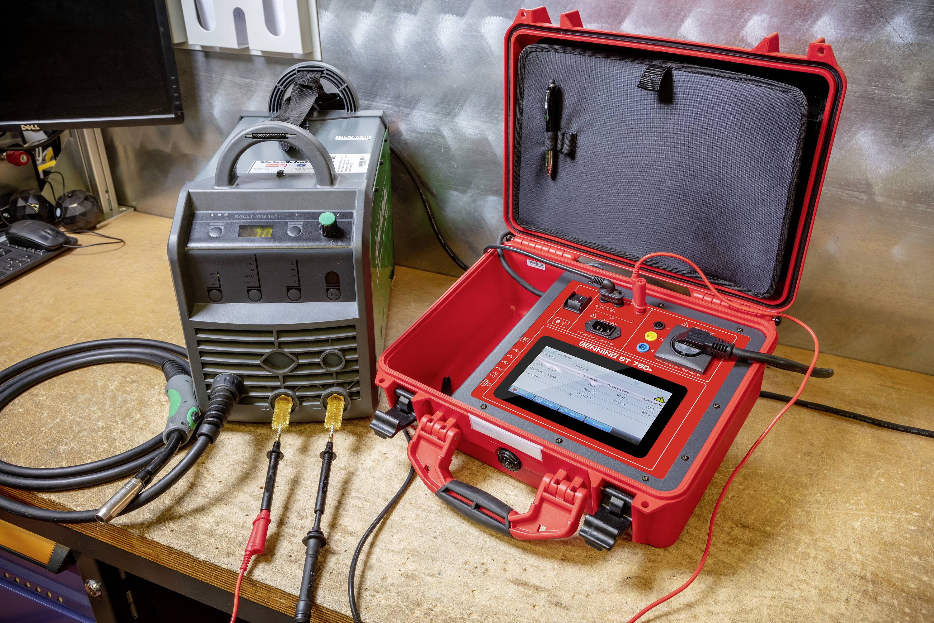 Een rood elektronisch testapparaat dat via kabels is verbonden met een ander apparaat op een werkbank. Op de achtergrond bevindt zich een metalen wand.