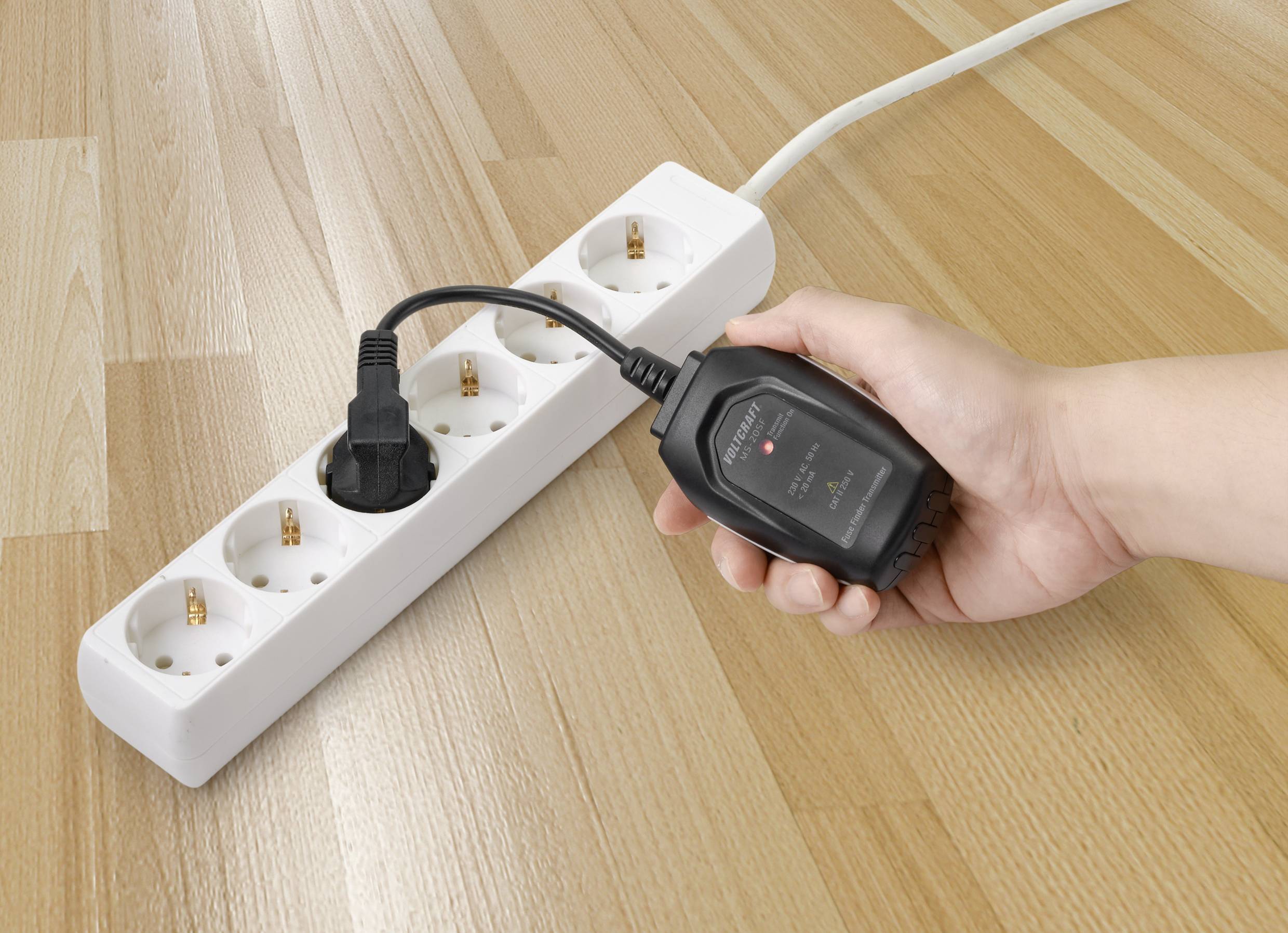 Een hand houdt een elektrische multimeter vast bij een stroomkabel die is aangesloten op een witte stekkerdoos om de stroomsterkte te controleren.