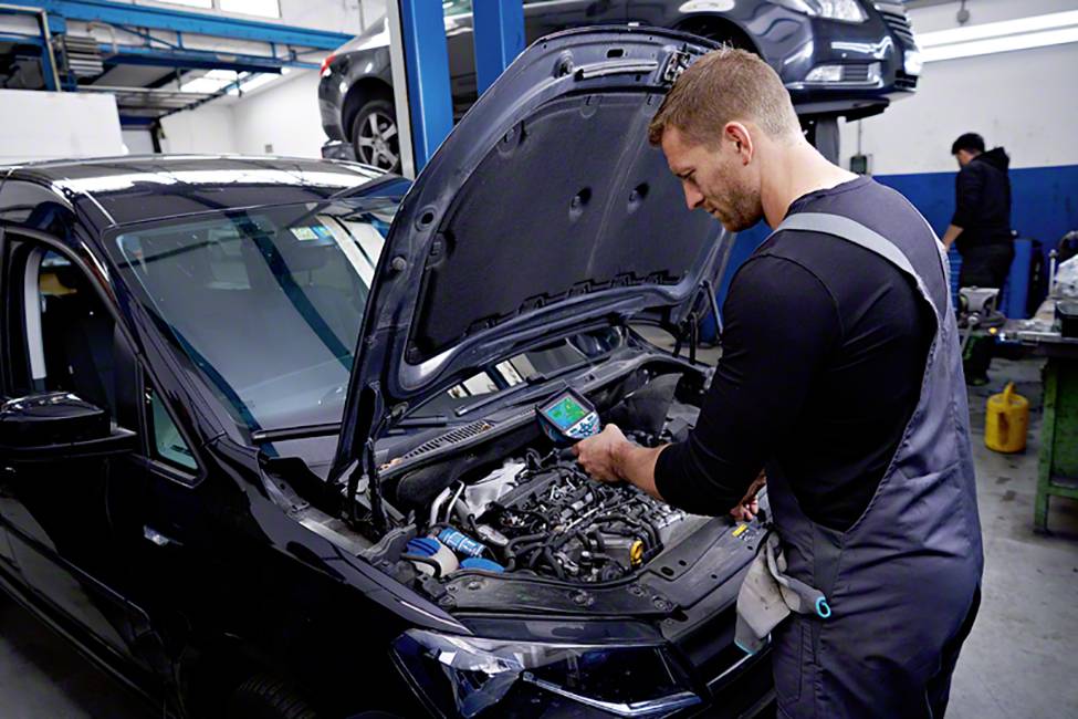 Een monteur controleert de motor van een zwarte auto in een garage met behulp van een diagnoseapparaat; andere auto's zijn op de achtergrond zichtbaar.
