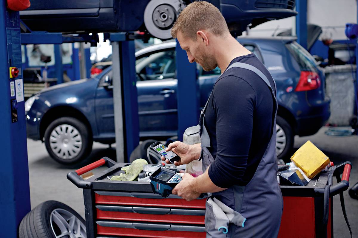 Een monteur in een werkplaats controleert een elektronisch apparaat naast een verrijdbare gereedschapswagen, terwijl een auto wordt opgetild.