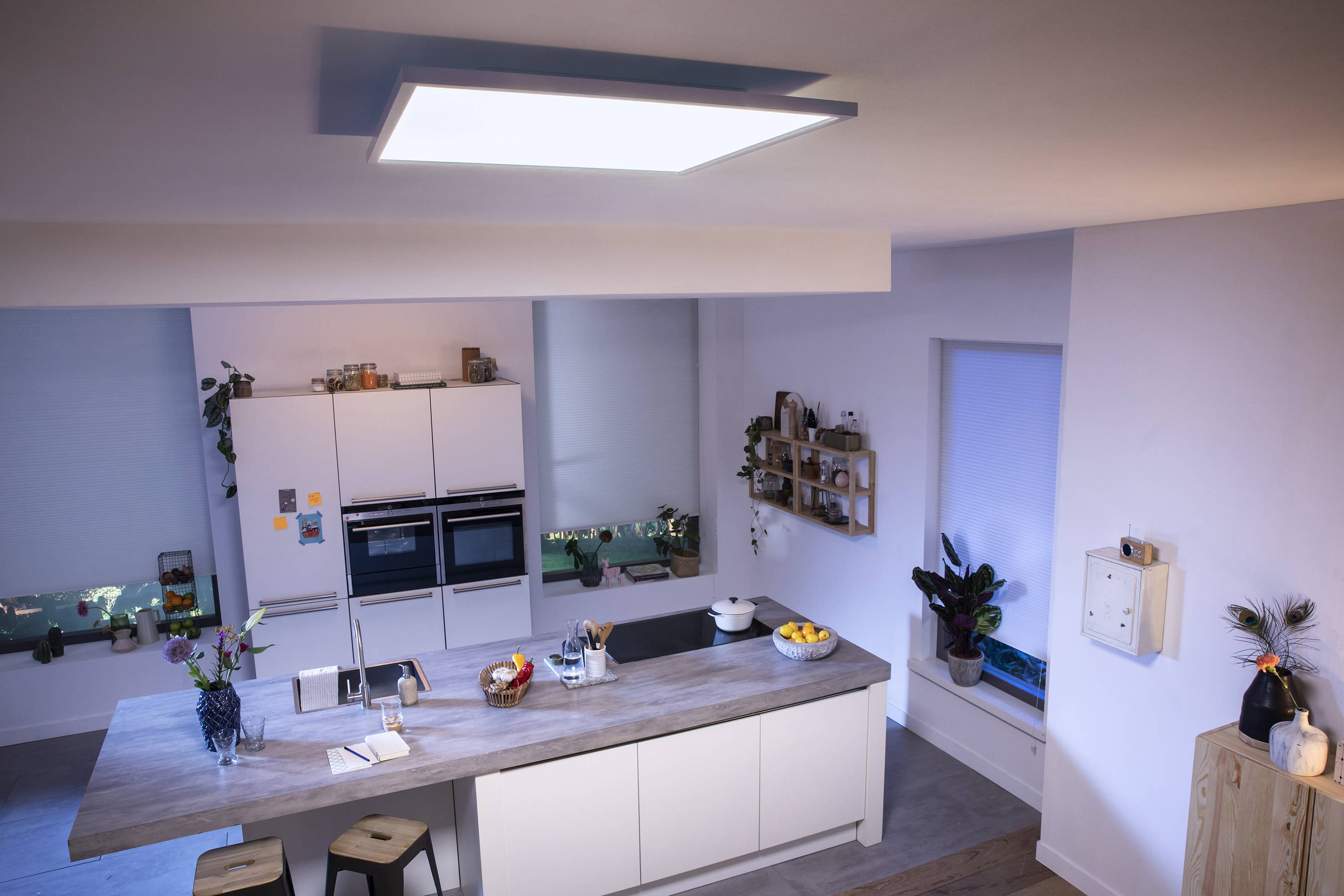 Moderne keuken met centrale houten toonbank, barkrukken en verlichting van bovenaf. Witte keukenkastjes aan de muur, planten als decoratie.