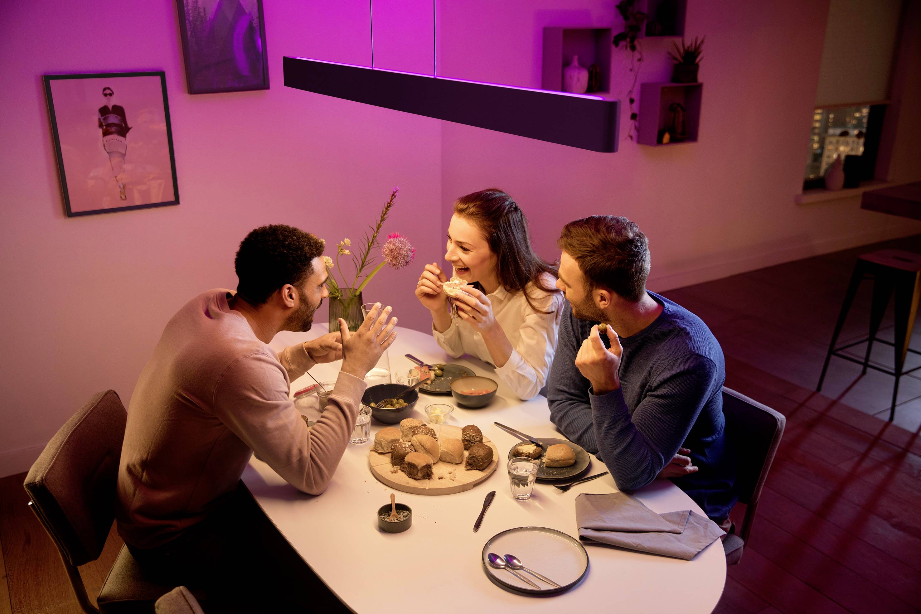 Drie personen zitten aan een tafel in een gezellige woonkamer en praten vrolijk terwijl ze eten. Een warme verlichting creëert een uitnodigende sfeer.