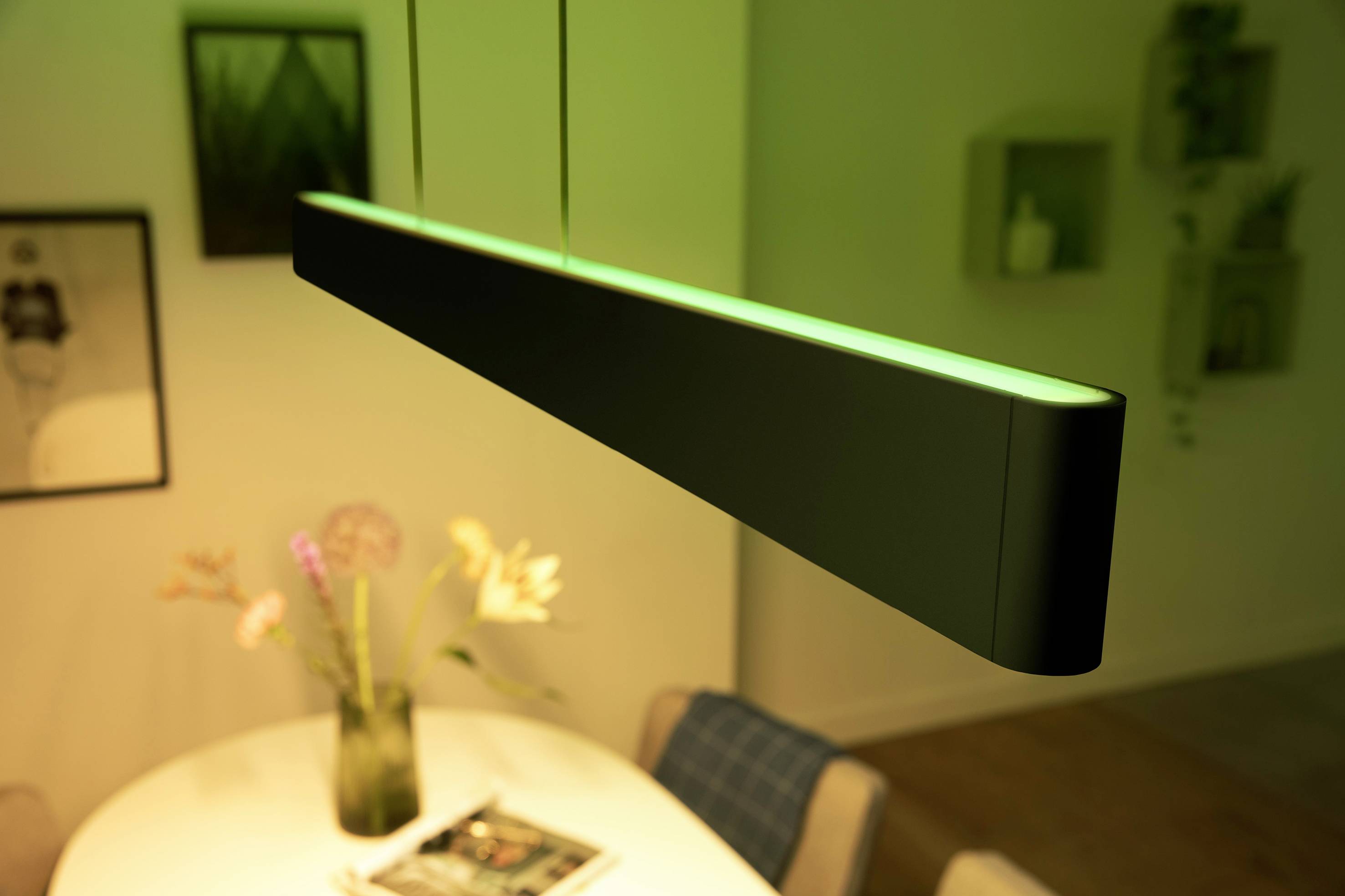 Hanglamp met groen licht boven een ronde tafel. Op de achtergrond zijn onscherpe fotolijsten en planken aan de muur zichtbaar.