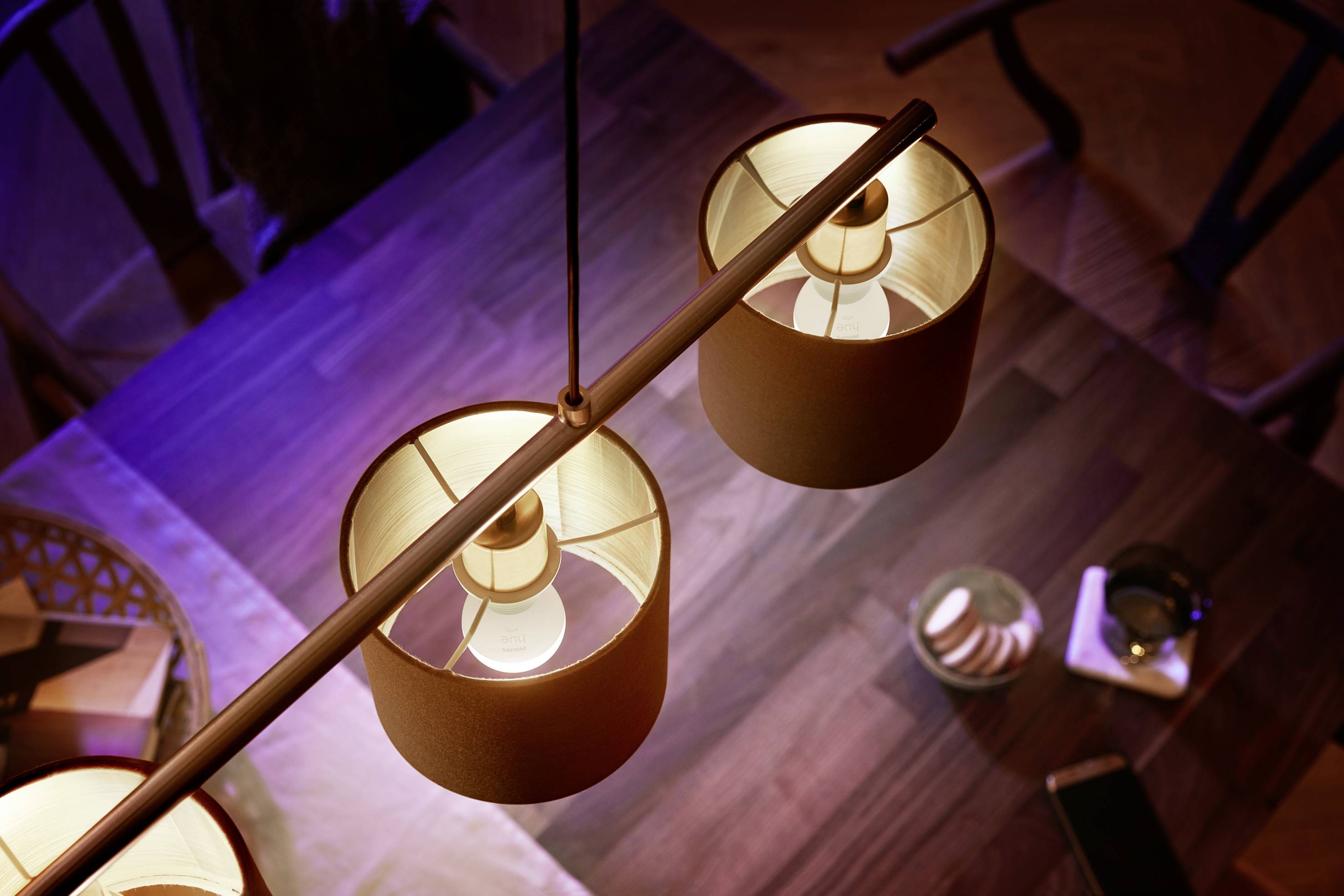 Twee hangende lampen schijnen boven een houten eettafel. Op de tafel staan kaarsen en een mobiele telefoon. Stoelen omringen de tafel.