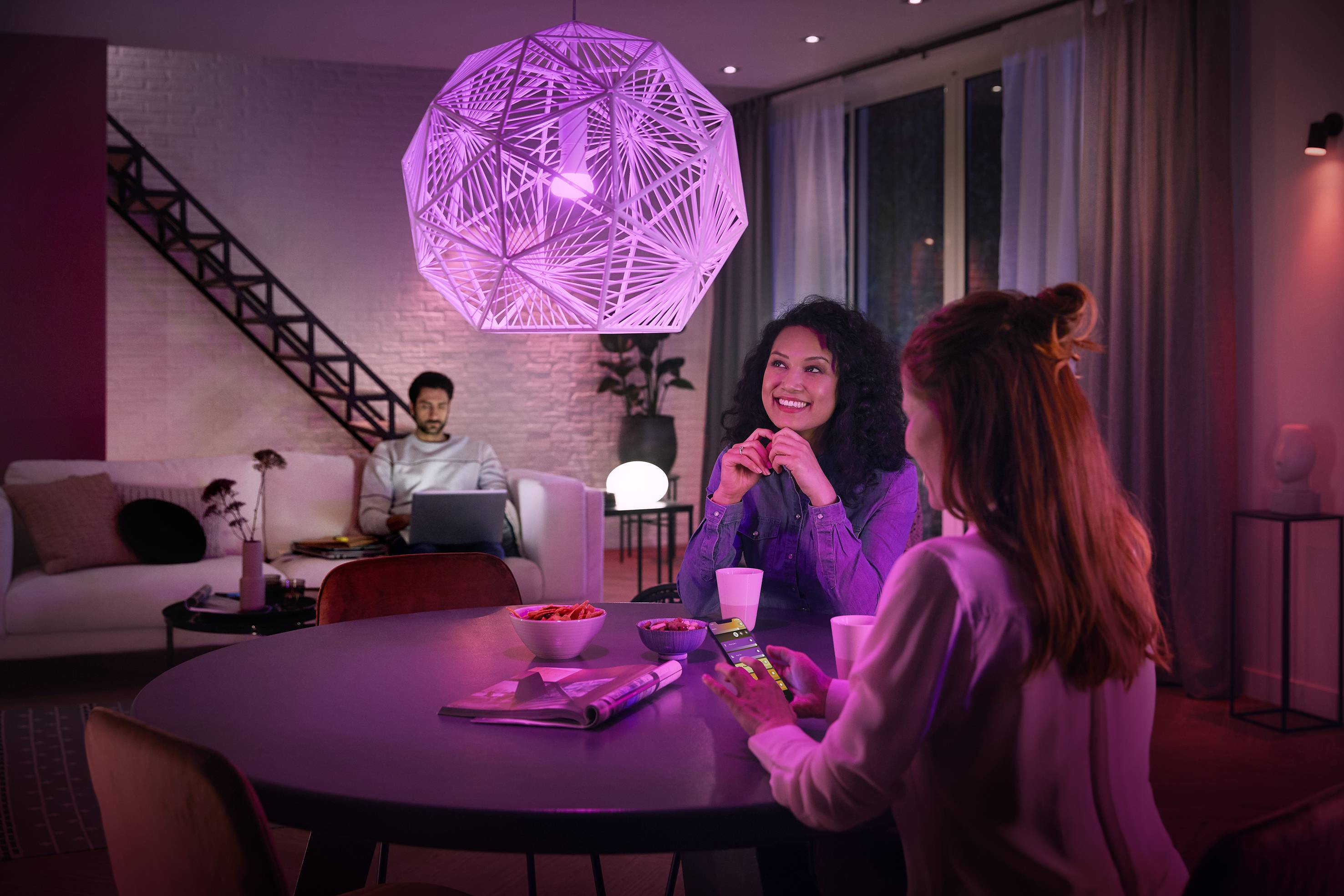 Een vrouw zit aan een tafel en praat vrolijk met een andere vrouw. Op de achtergrond werkt een man op een laptop.